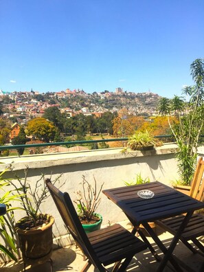 Un balcón con vistas a un paisaje urbano, que incluye una colina con edificios y una torre. Hay plantas en macetas y una mesa de madera con una silla.