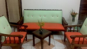 Una sala de estar con un sofá verde, dos sillas de madera, una mesita con un florero y un aparador de vidrio con libros.