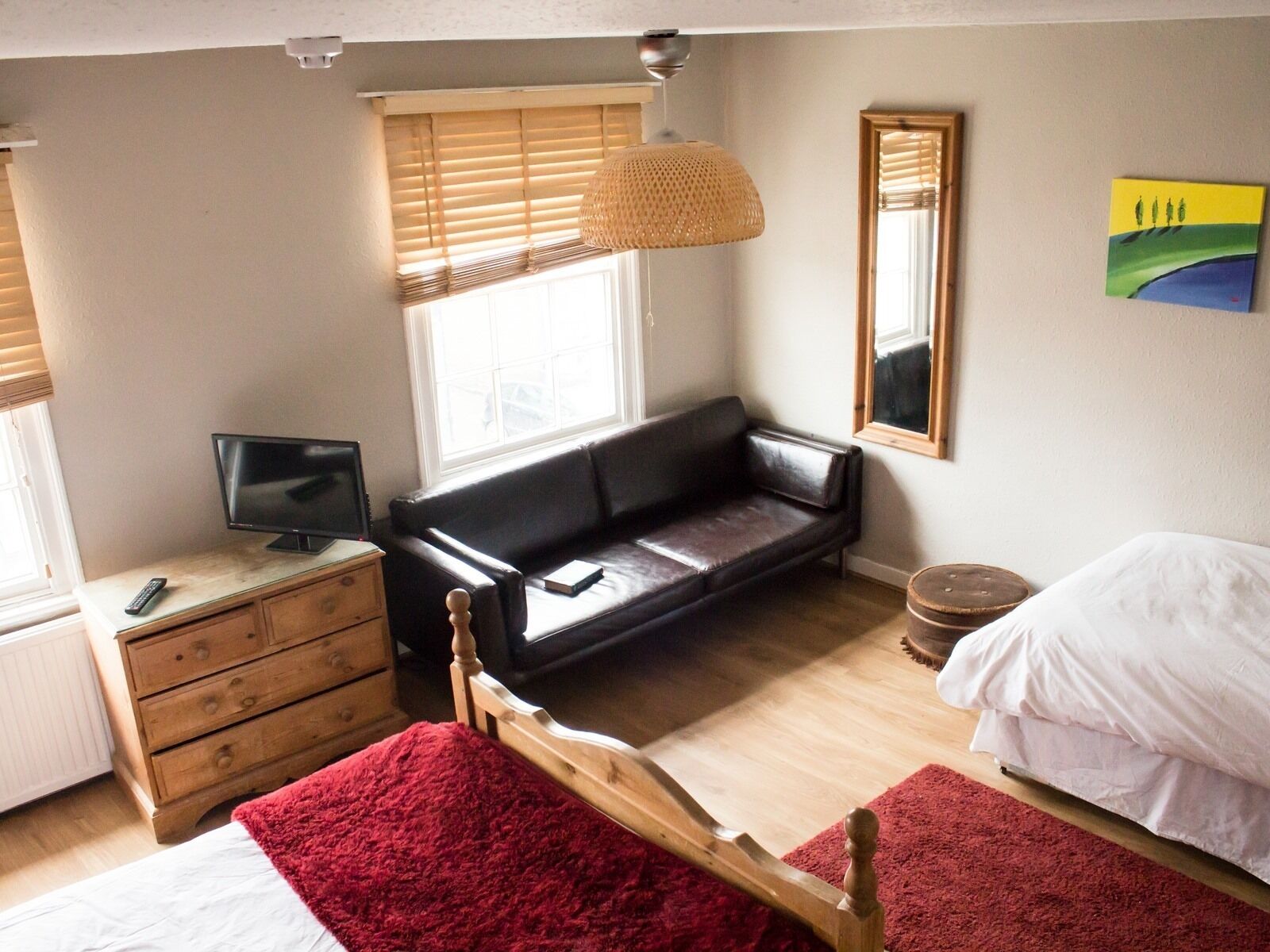 A bedroom with a bed, a wooden dresser, a black leather sofa, a television, a window with blinds, a mirror, and a painting on the wall.