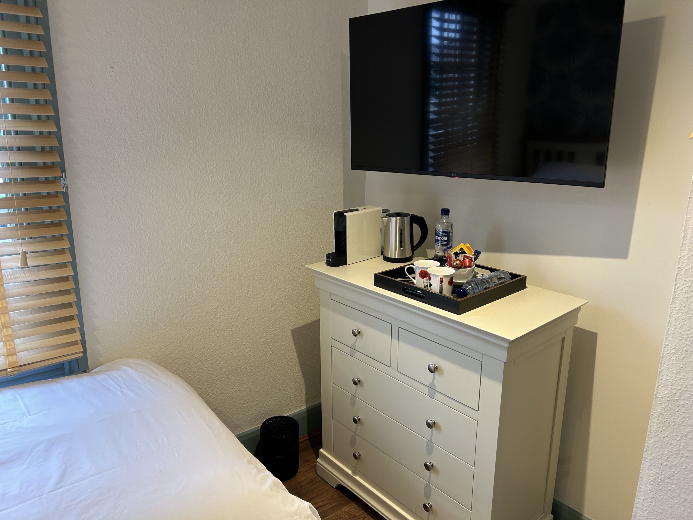 Ett hotellrum med en säng, en vit byrå med lådor, ett bricka med en kaffebryggare, en flaska vatten och en tv som är monterad på väggen.