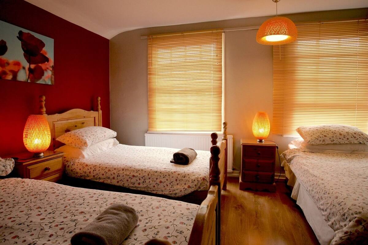 A bedroom with two single beds, a wooden headboard, a nightstand with a lamp, and a window with blinds.