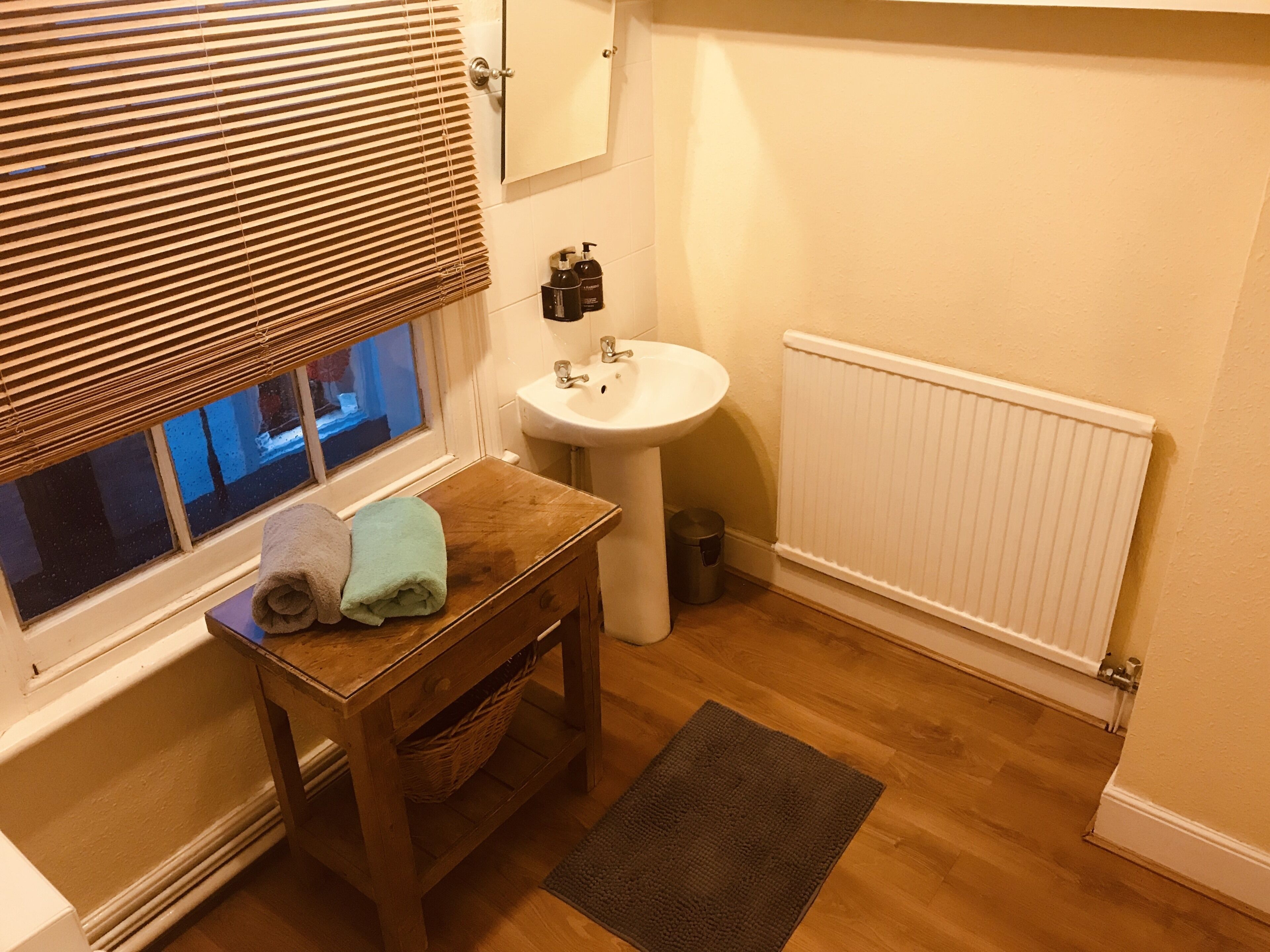 A bathroom with a wooden vanity, a sink, a window with blinds, a radiator, and a small rug on the floor.