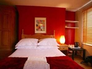 A bed with white linens, a red wall, a framed picture, a bedside lamp, and a small table with a cup. - The Ship (Chelmsford)