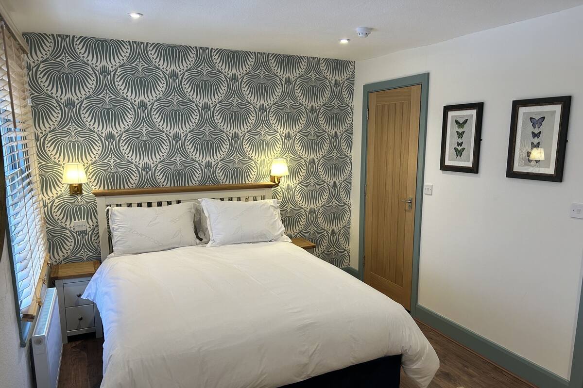 A neatly made bed with white linens, a headboard with a light wood finish, and a patterned wallpaper with a leaf-like design.