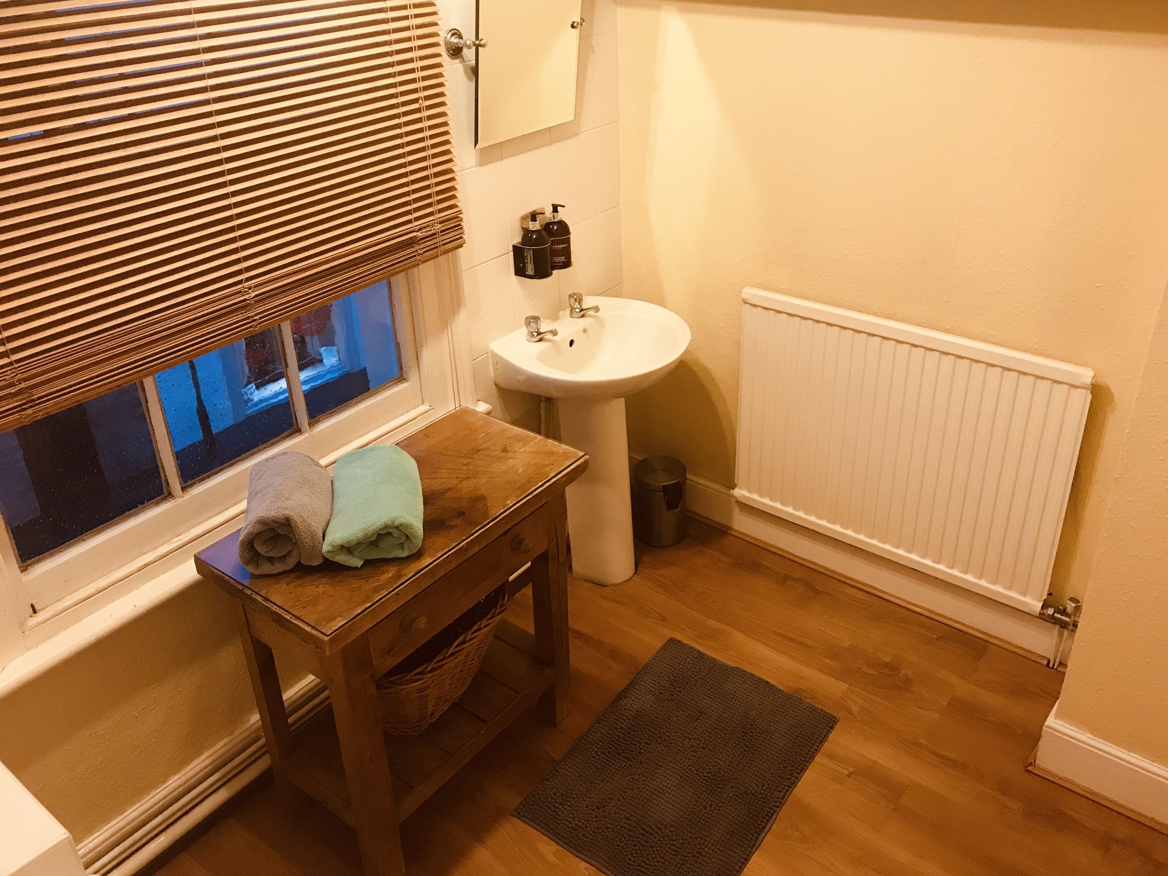 A bathroom with a wooden table, a sink, a window with blinds, and a radiator.