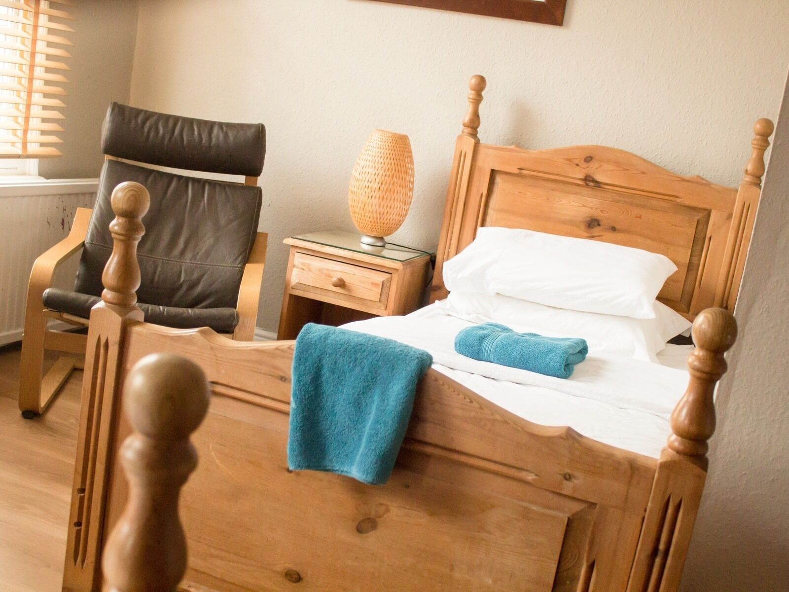 A wooden bed with white linens and a blue towel, a wooden nightstand with a lamp, and a leather chair.