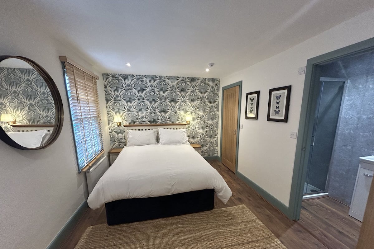 A bedroom with a bed, a round mirror, a patterned wallpaper, and a door leading to a bathroom.