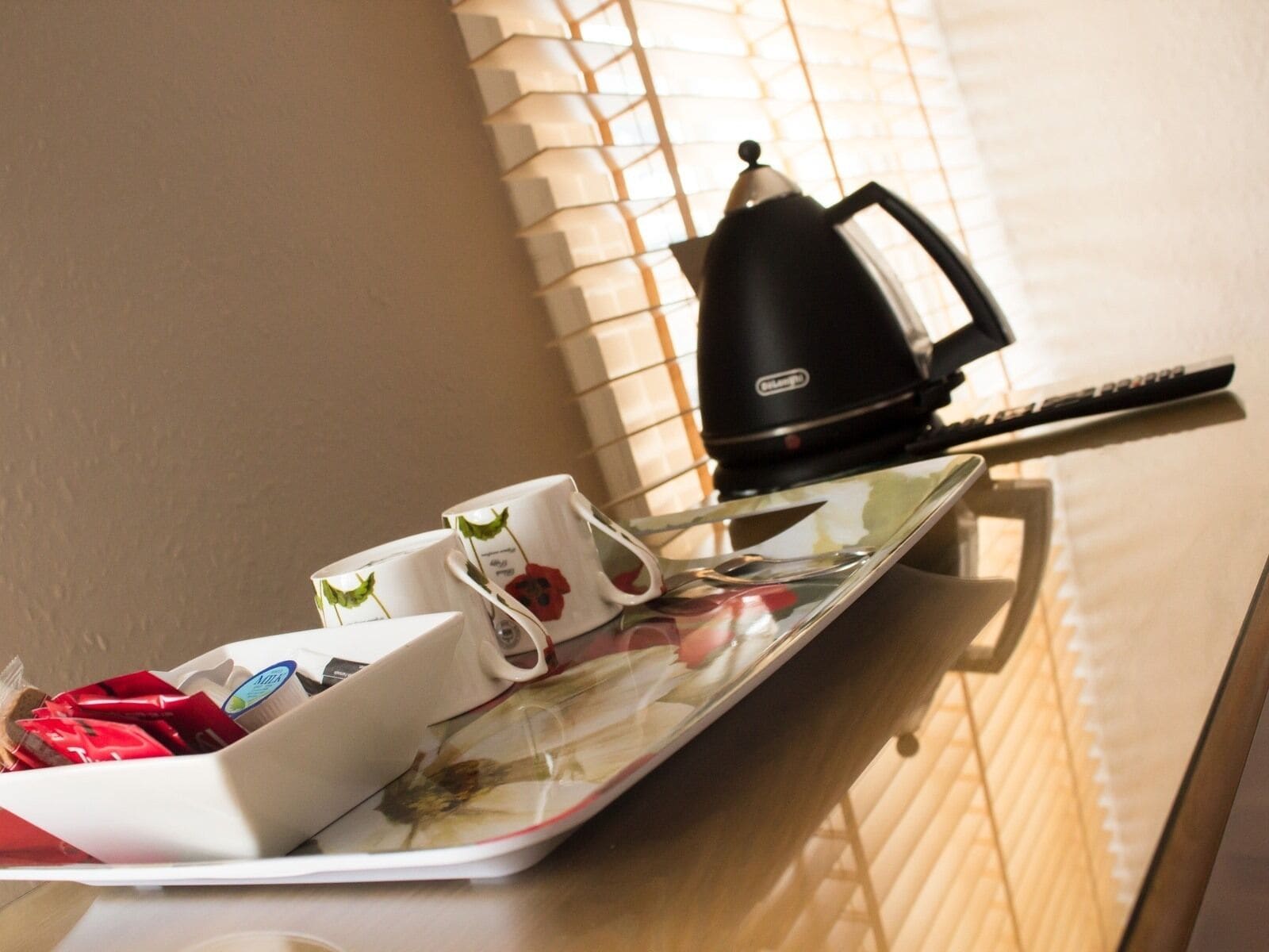 Un plateau avec une théière, des tasses et une petite assiette, posé sur une table près d’une fenêtre munie de stores.