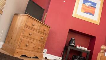 A room with a wooden dresser, a television on top, a framed picture on the wall, a small table with a kettle, and a trash bin.