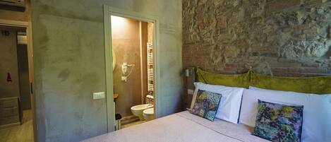 Un dormitorio con una cama grande, una pared de ladrillo, un baño con lavamanos y una puerta que da a otra habitación.