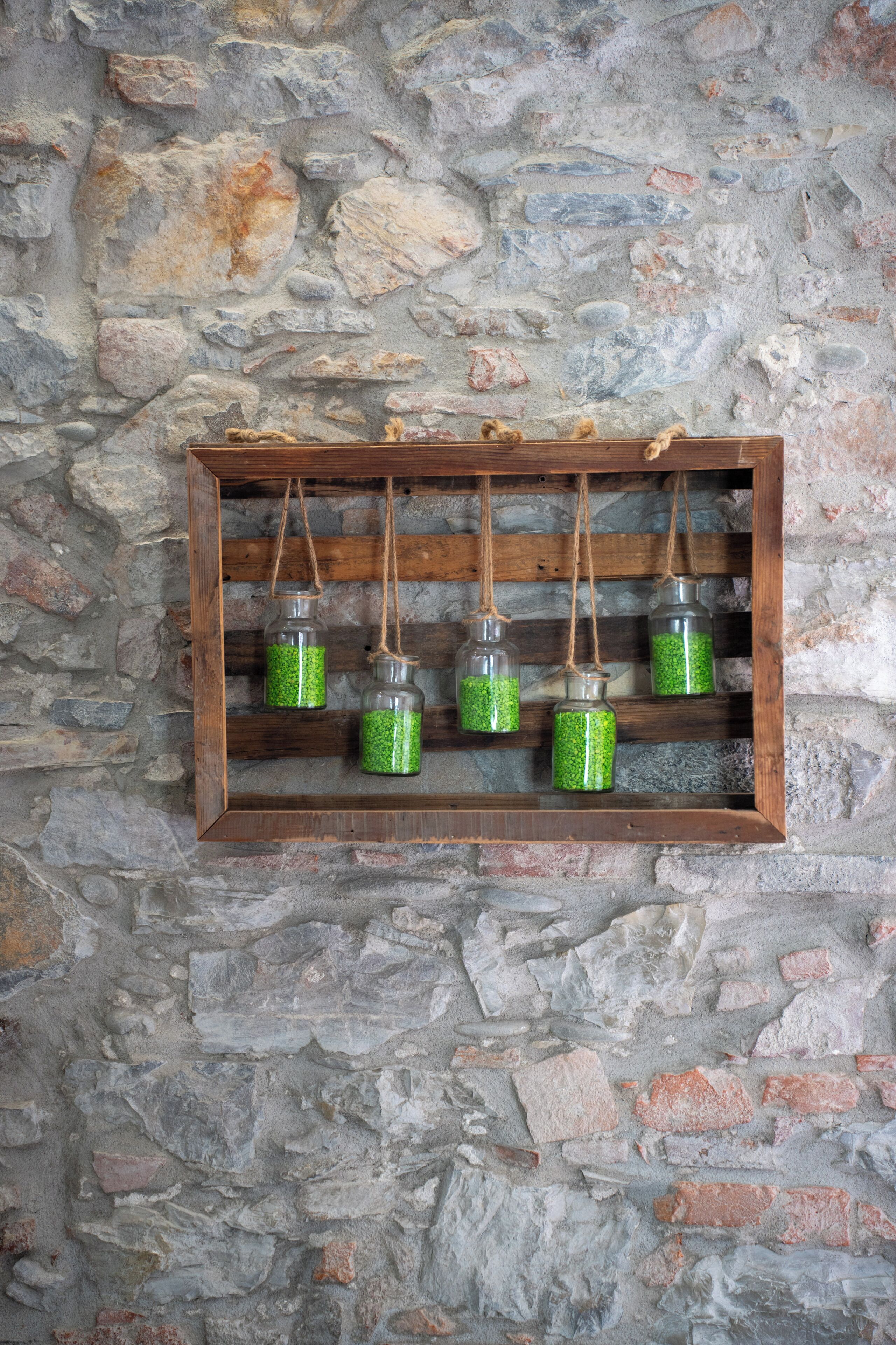 A wooden shelf with four hanging glass jars against a stone wall.