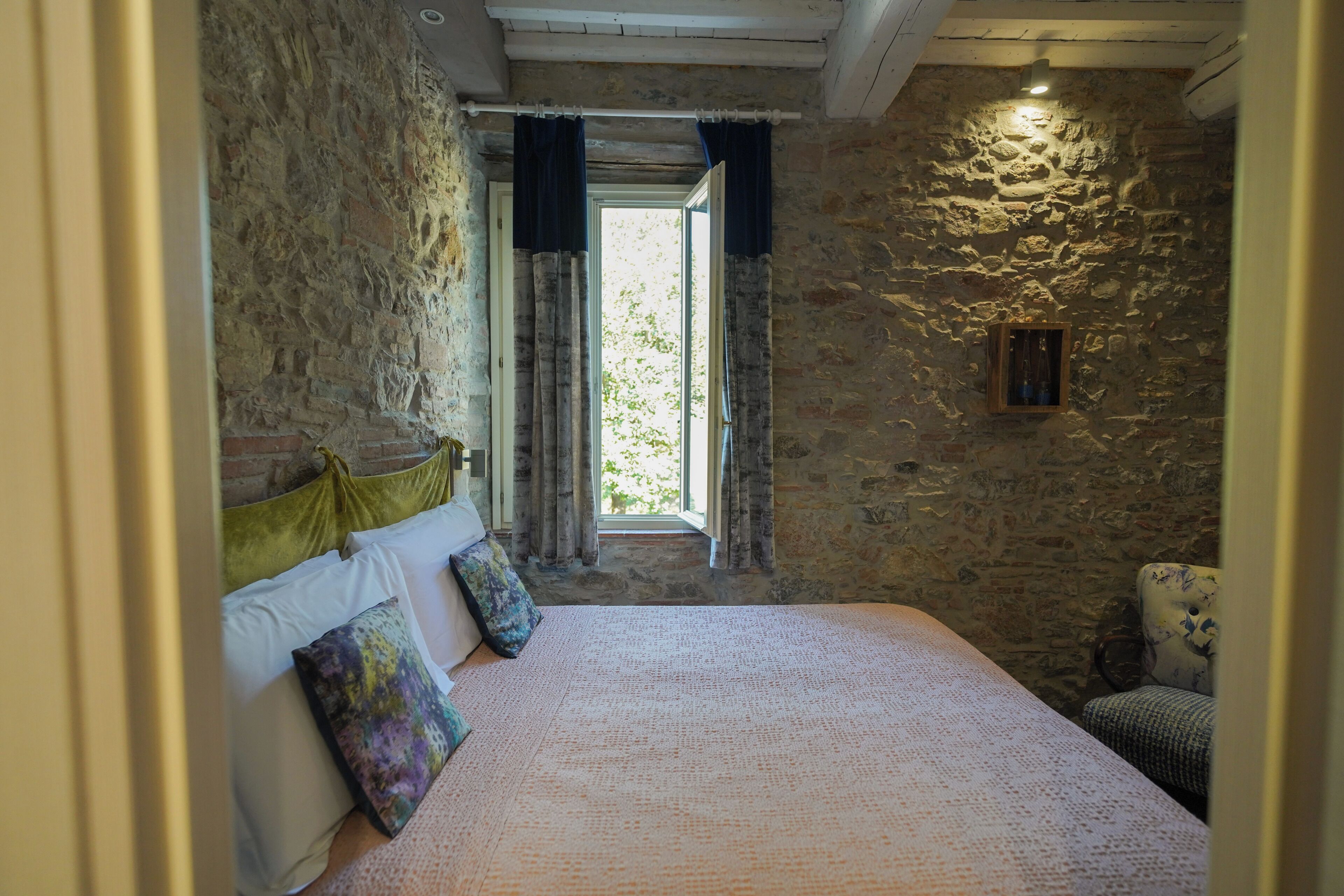 Un dormitorio con pared de piedra, cama con cubrecama estampado, ventana con cortinas y un marco de madera sencillo.