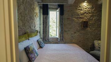 A bedroom with a stone wall, a bed with a patterned bedspread, a window with curtains, and a small wooden window frame.