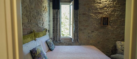 Un dormitorio con pared de piedra, cama con cubrecama estampado, ventana con cortinas y un marco de madera sencillo.