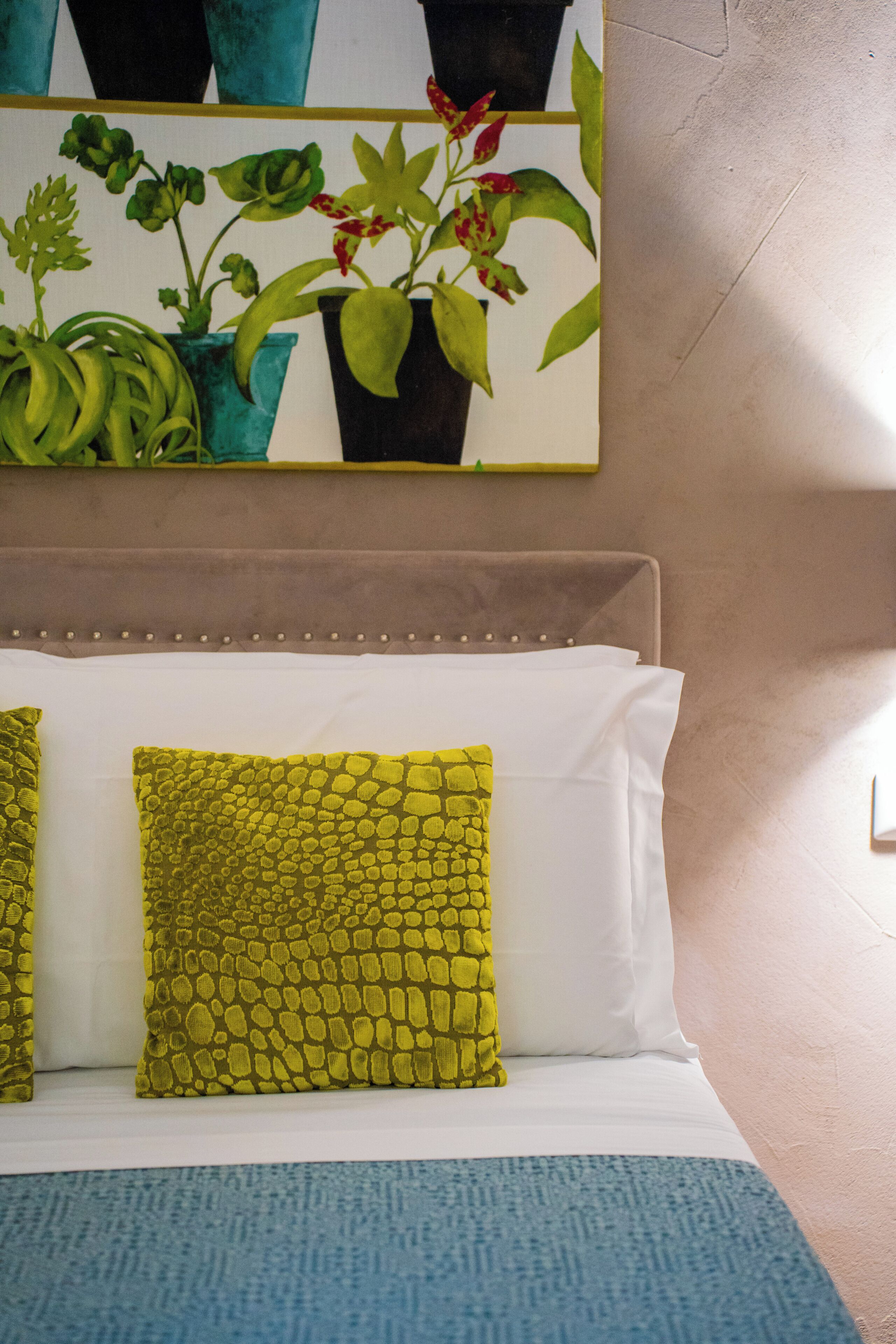 A bed with white and green pillows, a blue throw blanket, and a painting of plants on the wall.