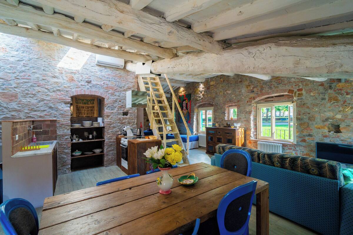A rustic interior with a stone wall, wooden furniture, a staircase, and a view of the outdoors.