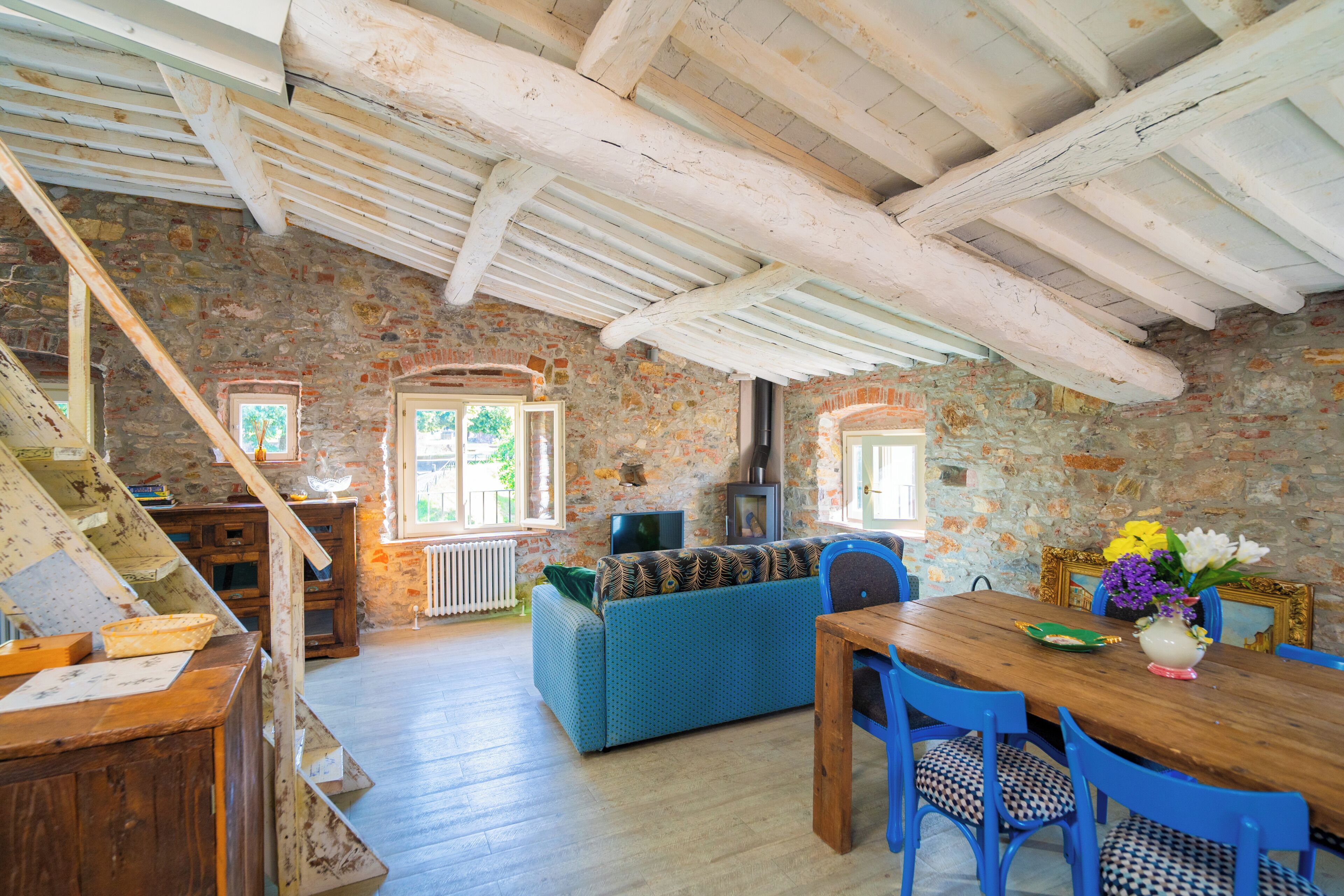 A rustic interior with exposed wooden beams, a stone wall, a blue sofa, and a wooden dining table with chairs.