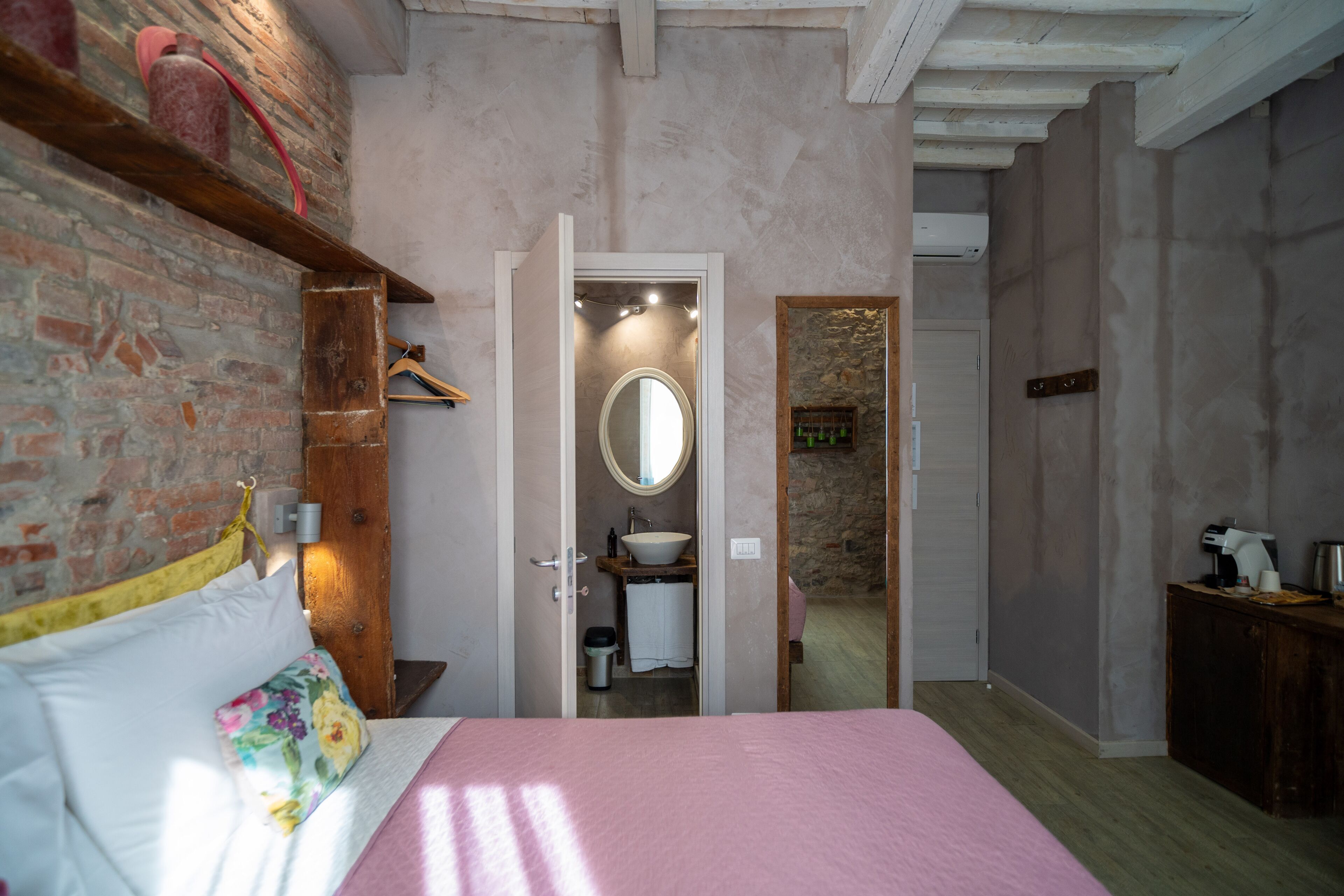 Een slaapkamer met een bed, een houten hoofdeinde, een bakstenen muur, een spiegel en een badkamer zichtbaar door een open deur.