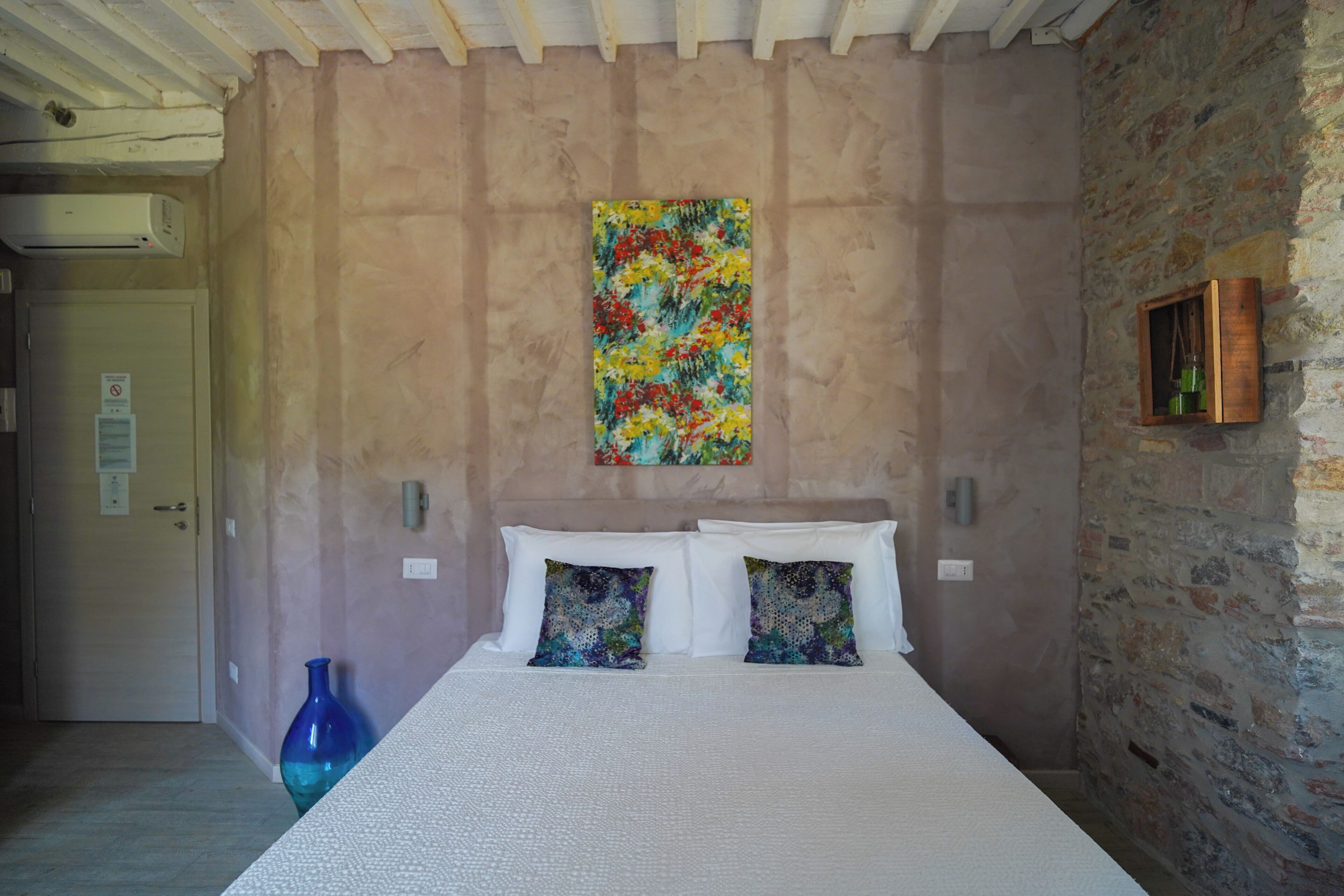 A bedroom with a bed, two pillows, a colorful painting, a blue vase, and a stone wall.
