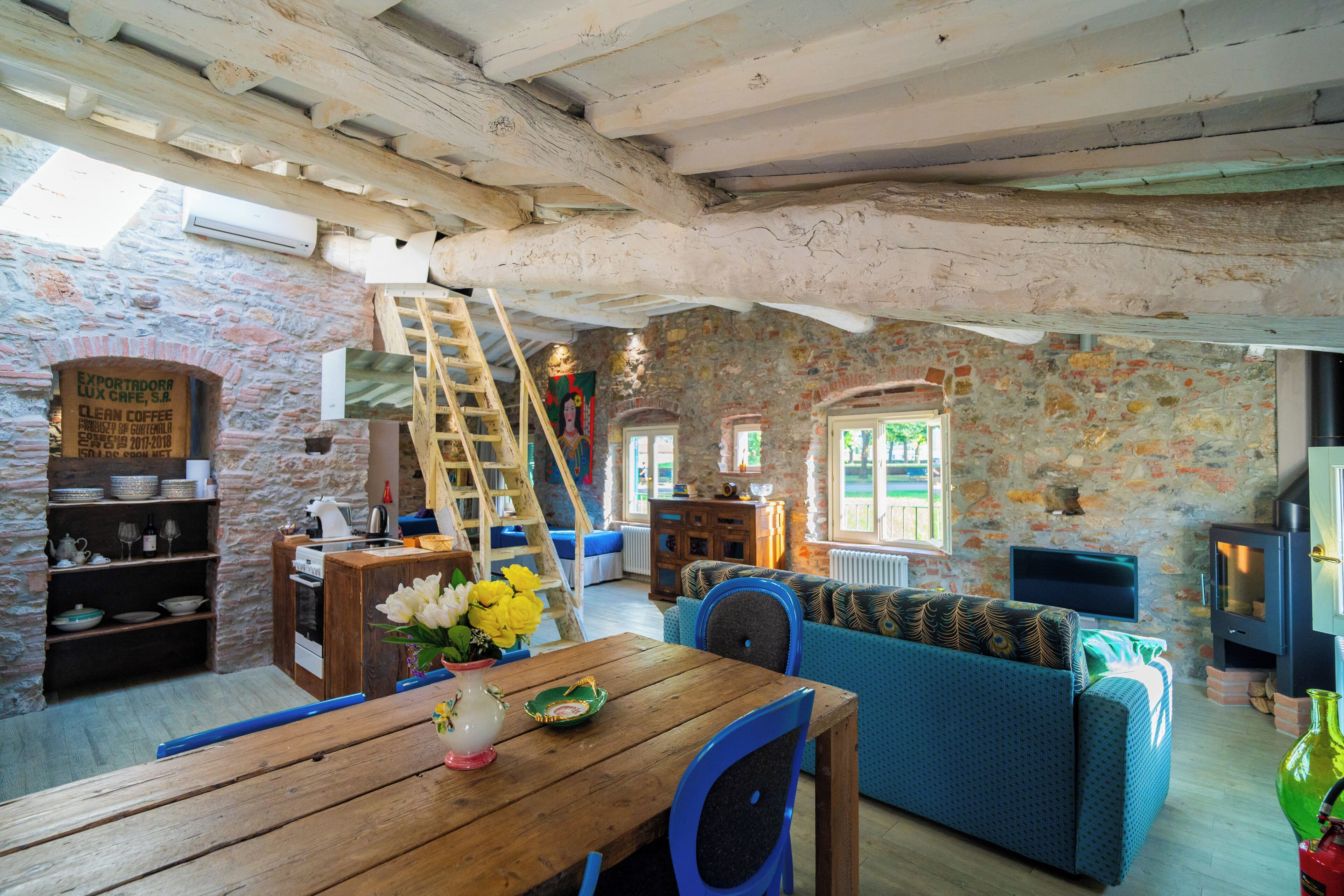 A rustic interior with exposed wooden beams, a stone wall, a wooden table with a vase, a blue sofa, and a television.