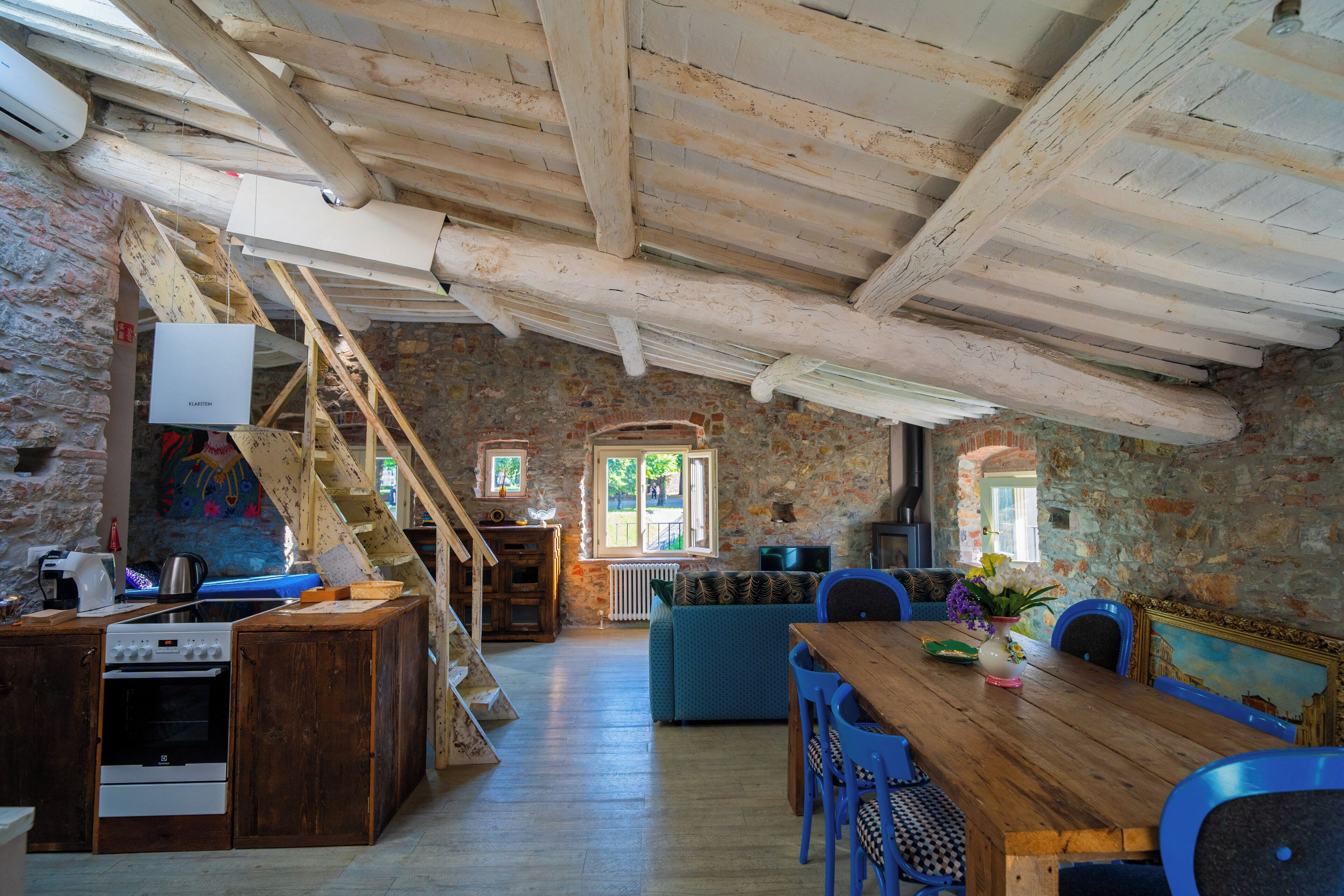 A rustic kitchen with a wooden stove, a dining table with chairs, and a stone wall.