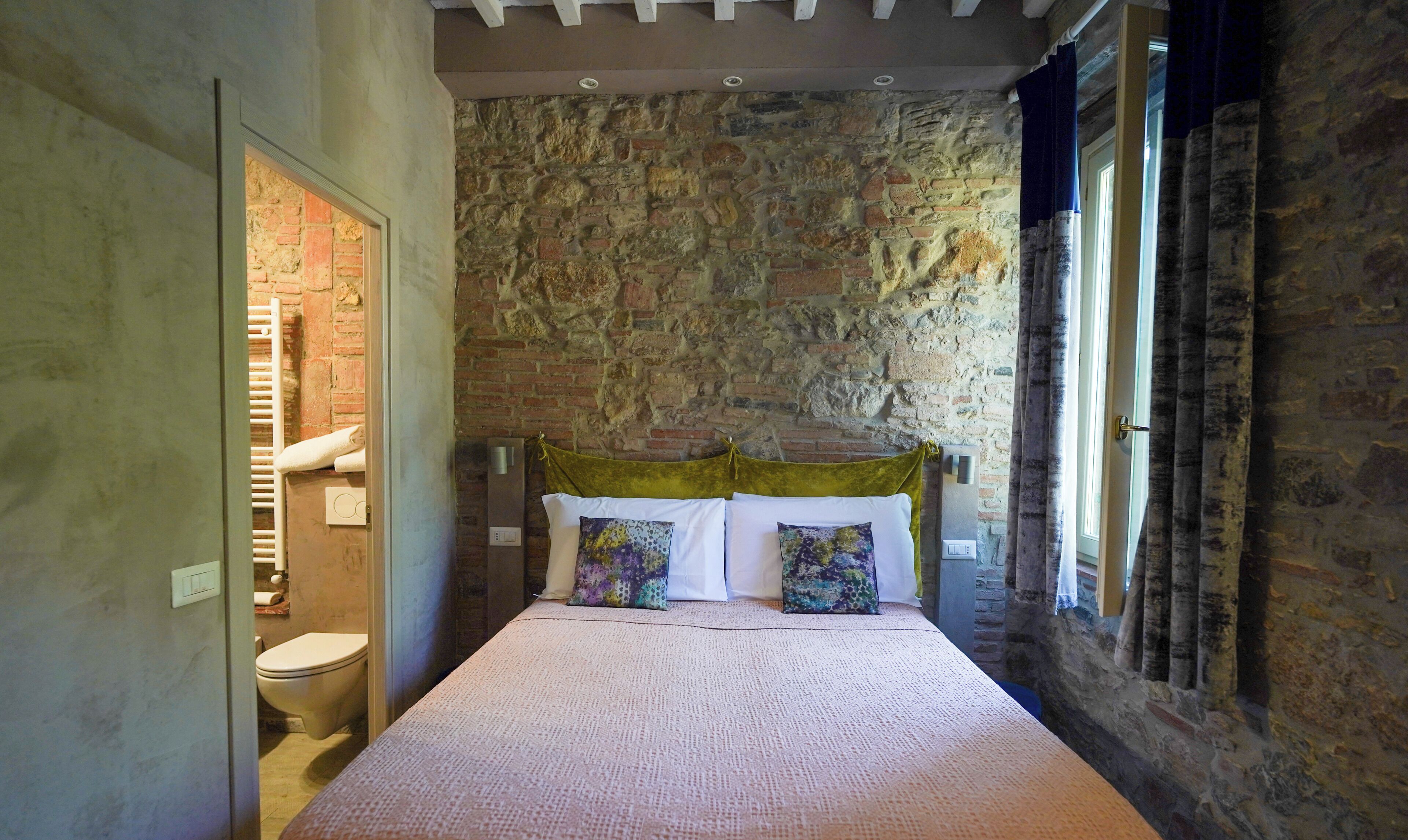A bedroom with a stone wall, a bed with patterned pillows, a window with curtains, and a visible bathroom.