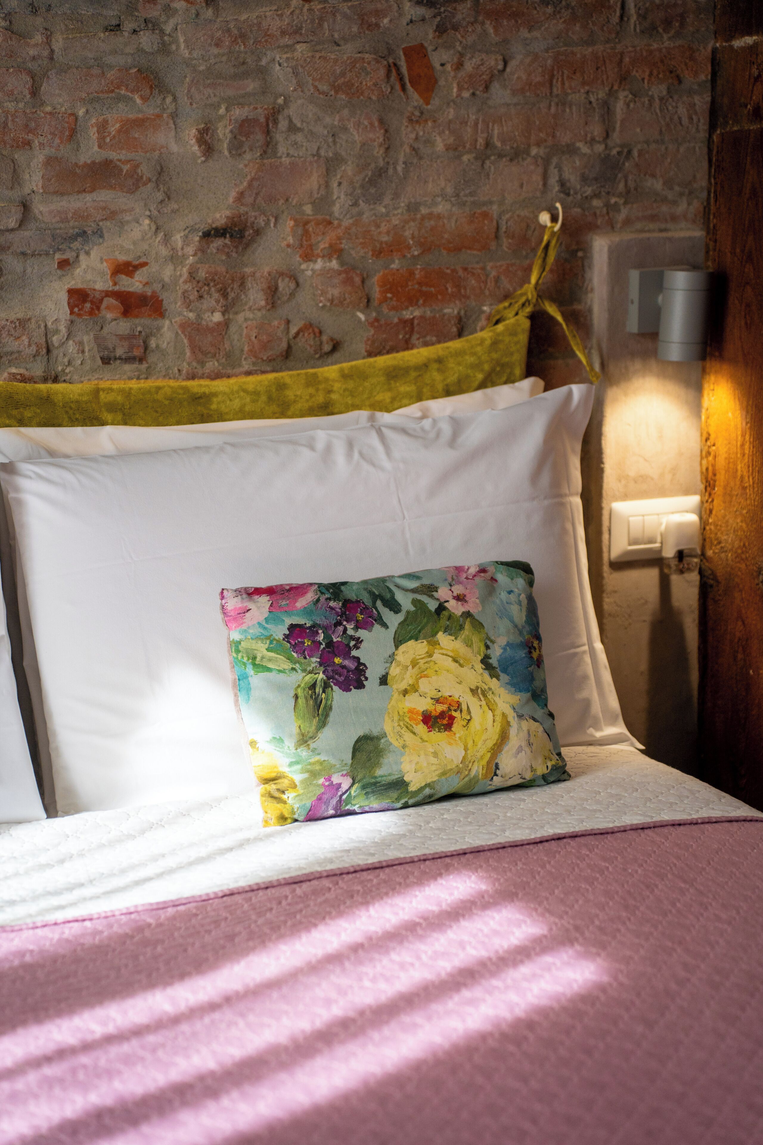A bed with white pillows and a floral pillow, a brick wall, and a wall-mounted light fixture.