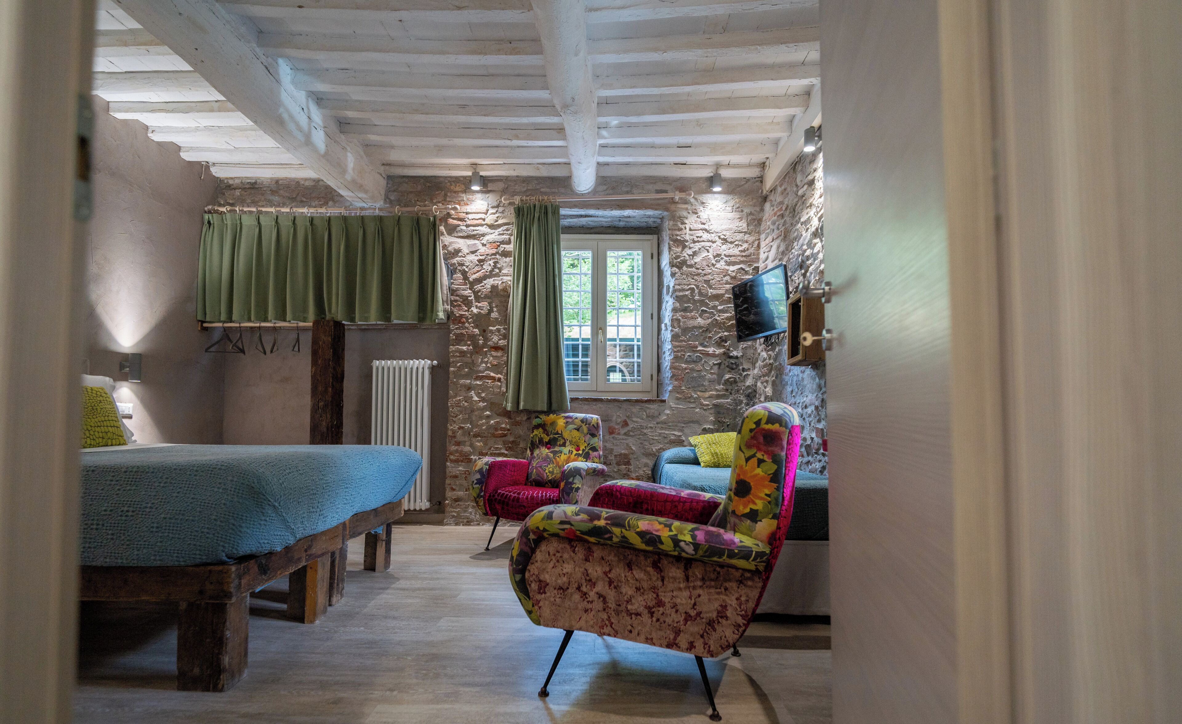 A rustic room with a bed, a chair, a radiator, a window with curtains, and a television on the wall.