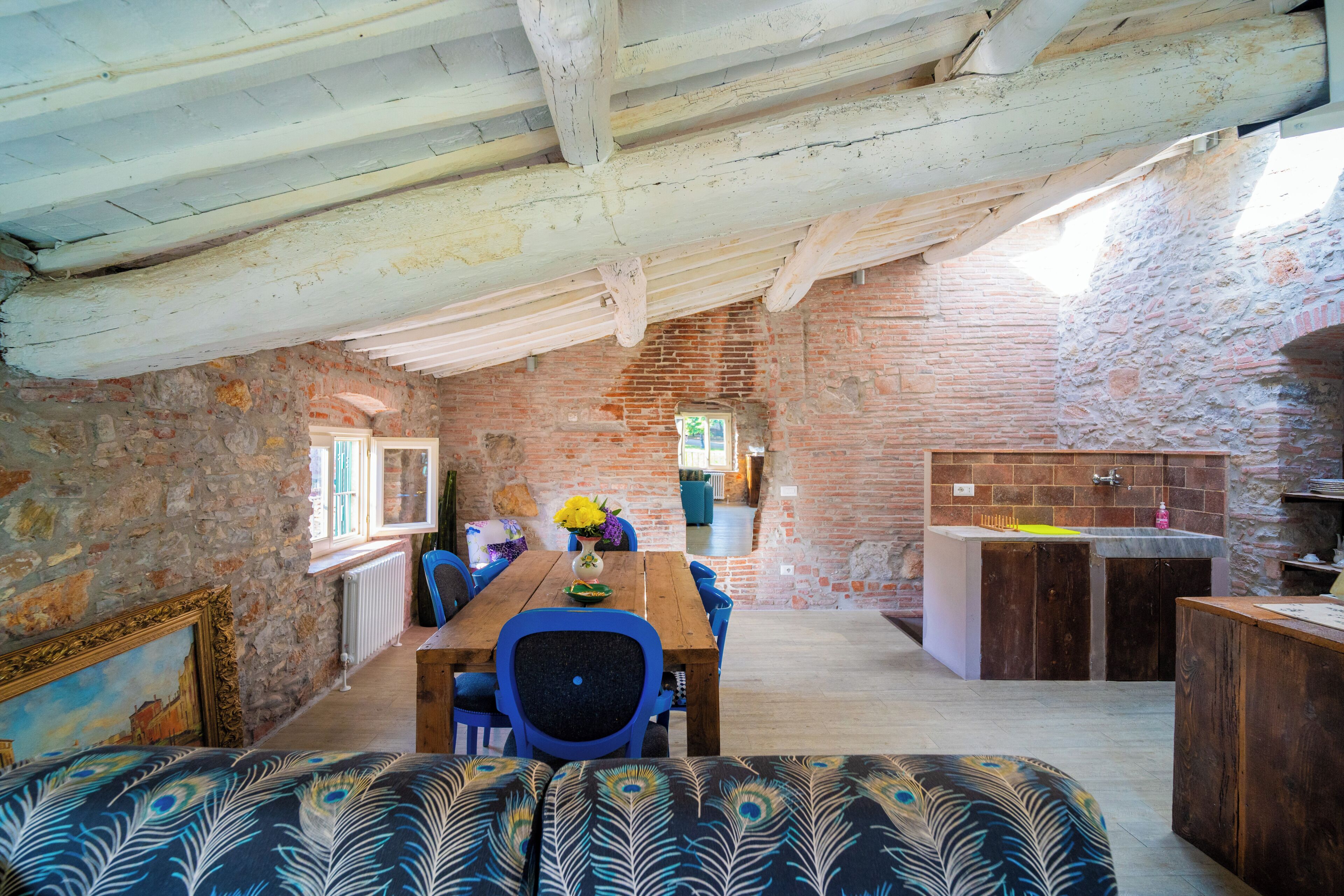 A rustic interior with exposed wooden beams, a dining area with a wooden table and chairs, a stone wall, and a peacock-patterned sofa.