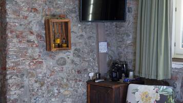 Une pièce avec un mur en pierre, un téléviseur fixé au-dessus d’un meuble en bois, un fauteuil à motifs floraux et un petit tabouret en bois.