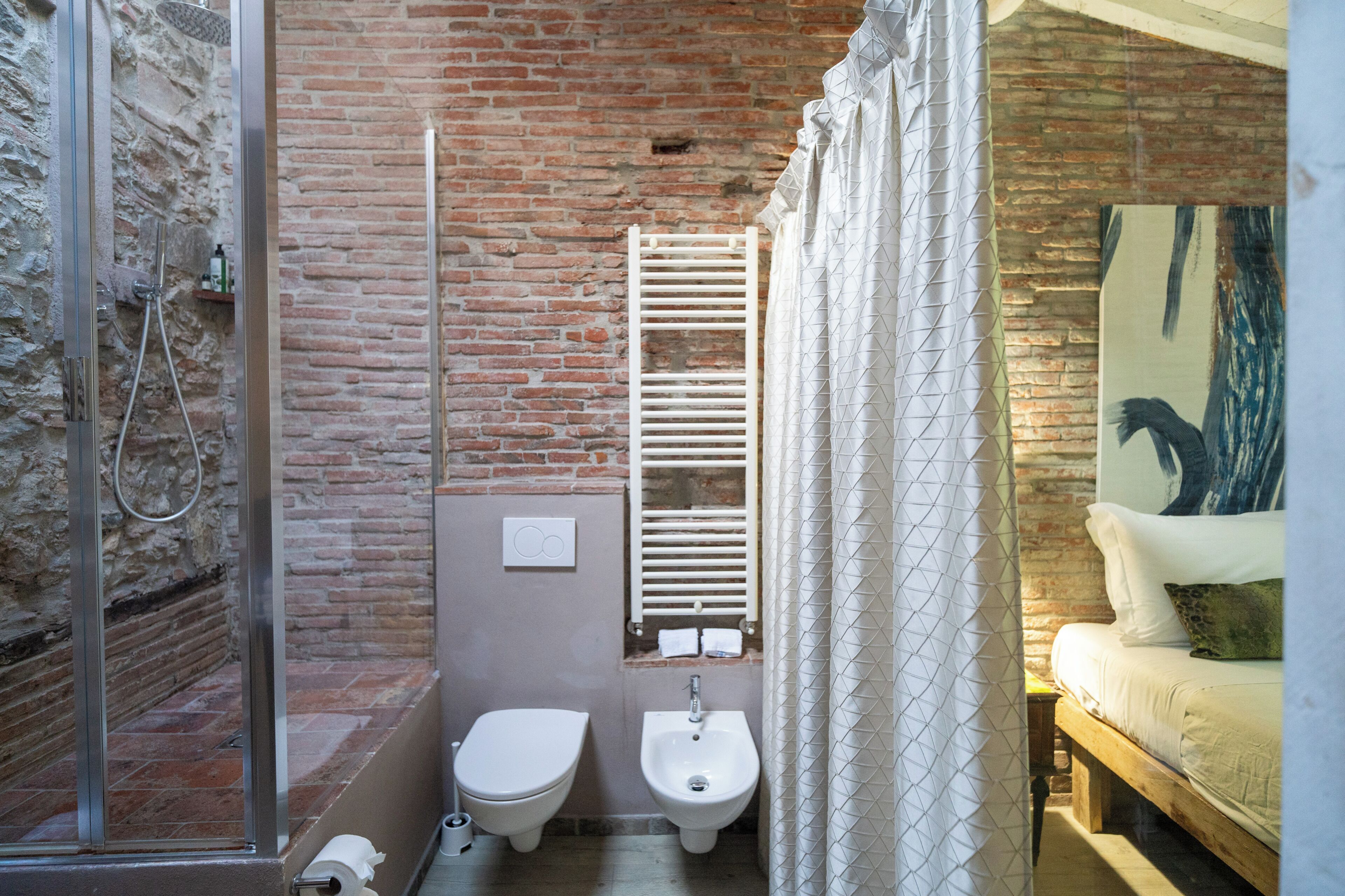 A bathroom with a shower, toilet, bidet, and a bed with a painting.