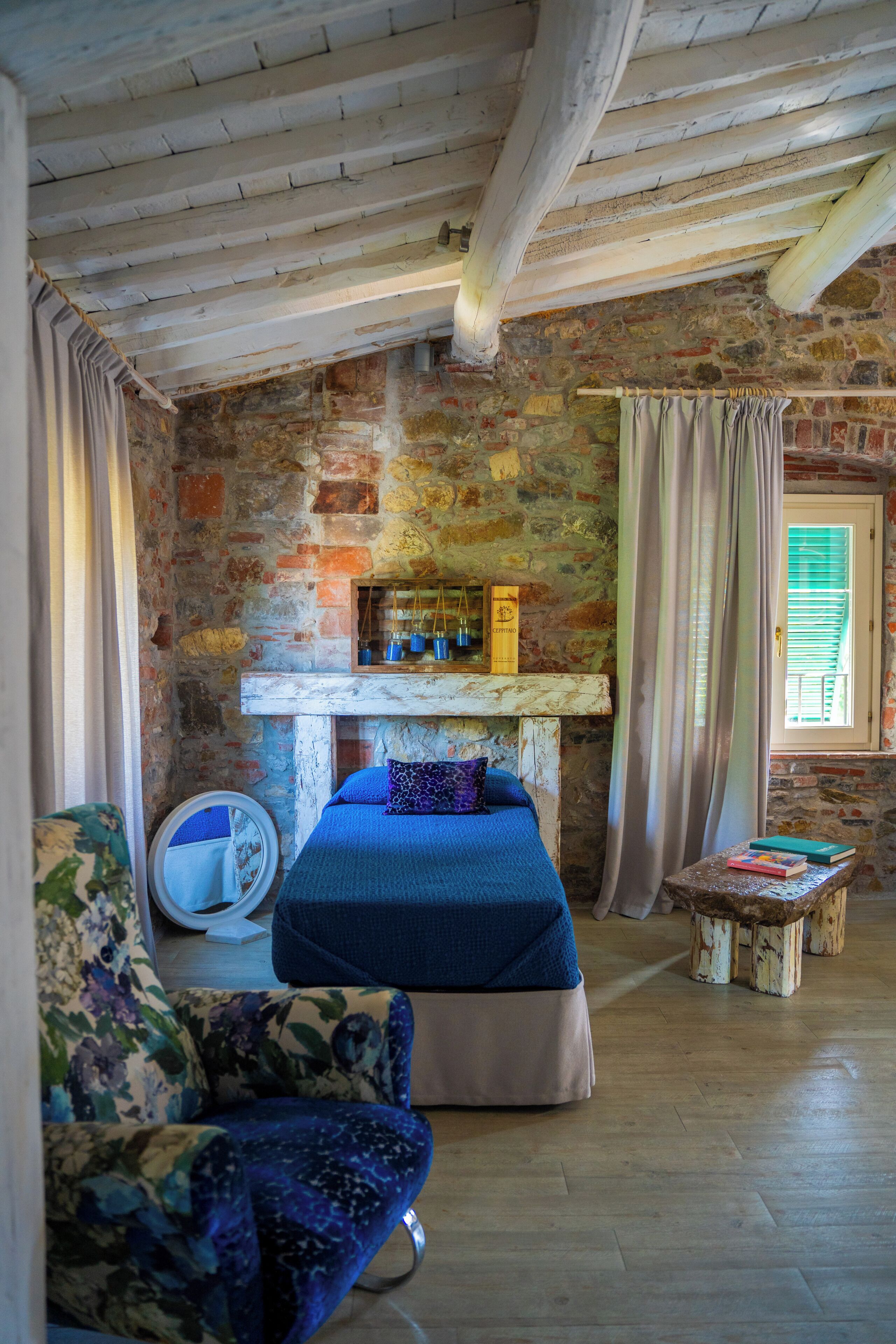 A rustic room with exposed brick walls, a wooden ceiling, a bed with blue bedding, a round mirror, and a small table with books.