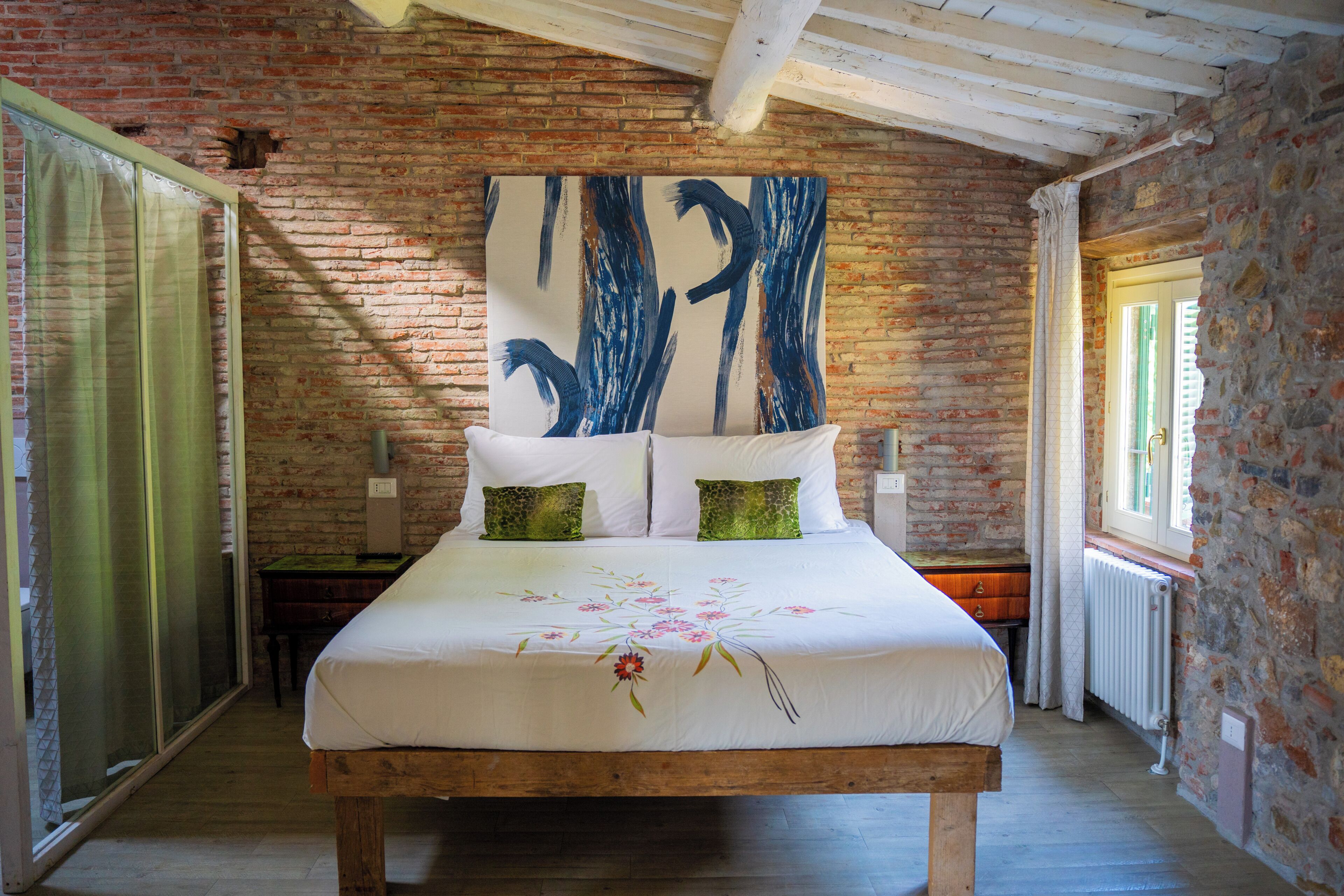 A bedroom with a brick wall, a large bed with white bedding, two green pillows, and a floral design.