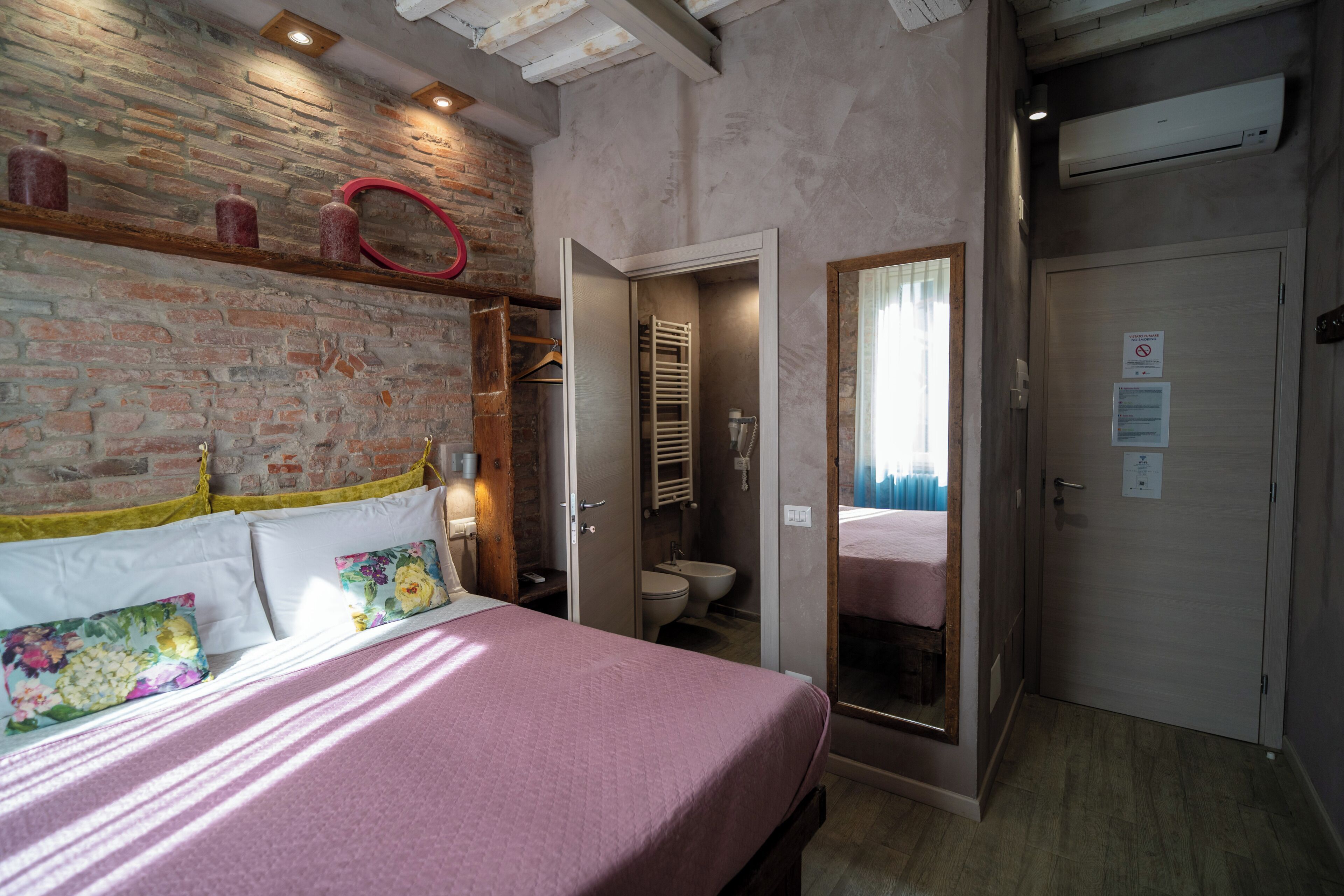 A bedroom with a brick wall, a bed with floral pillows, a wooden headboard, and a mirror.