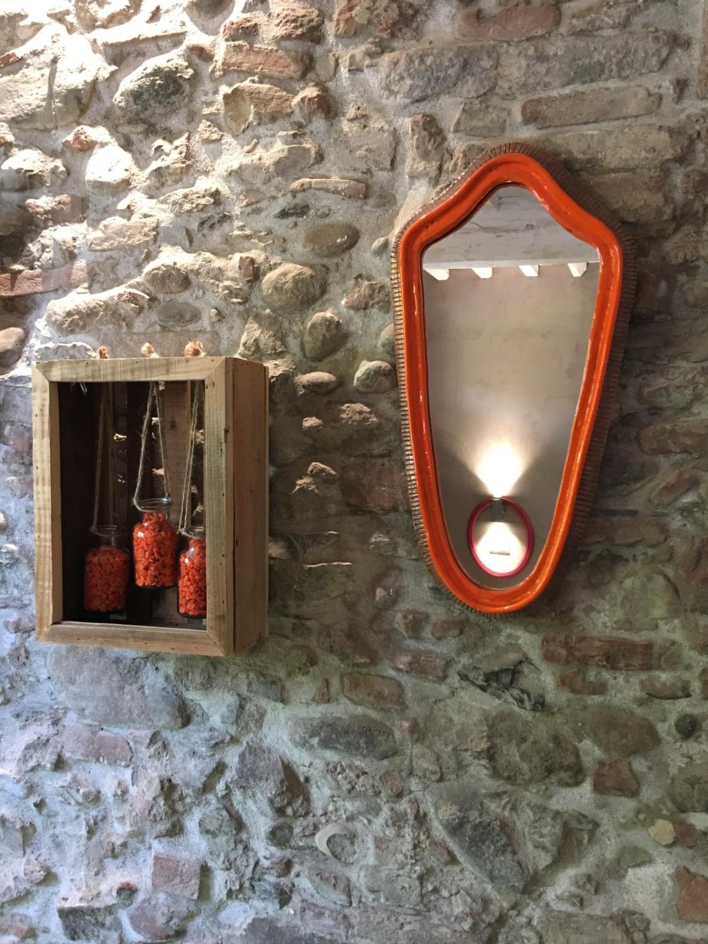 Un mur de pierres rustiques surmonté d’une boîte en bois contenant des bocaux en verre suspendus et d’un miroir avec un cadre orange.