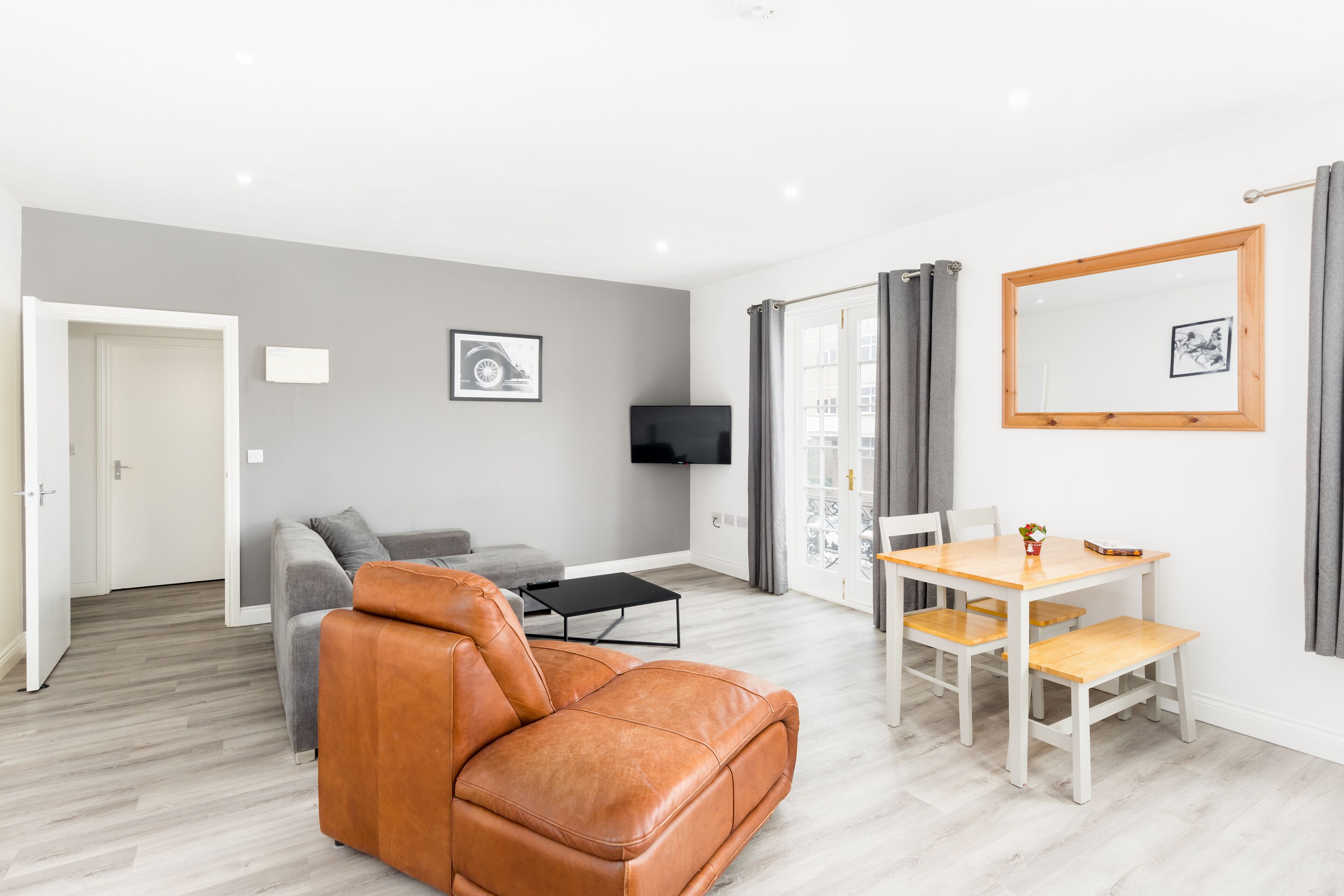 A modern living room with a brown leather sofa, a grey sofa, a wooden dining table with chairs, a flat-screen TV mounted on the wall, and a framed picture.