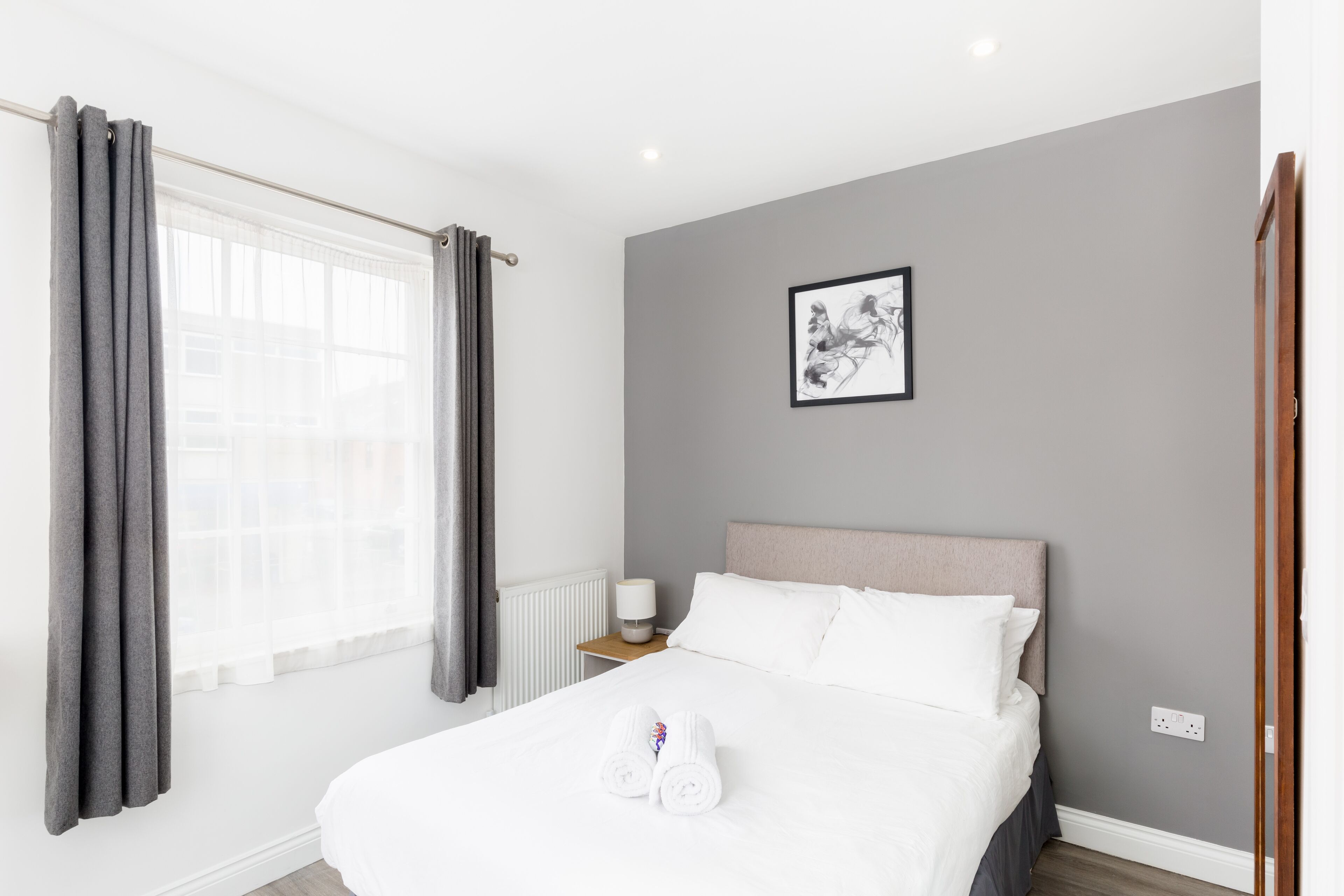 Una cama bien hecha con sábanas blancas y toallas enrolladas, una mesita de noche con una lámpara y una obra de arte enmarcada en la pared.