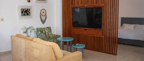 A living room with a sofa, two small tables, a TV, and a bed in the background.