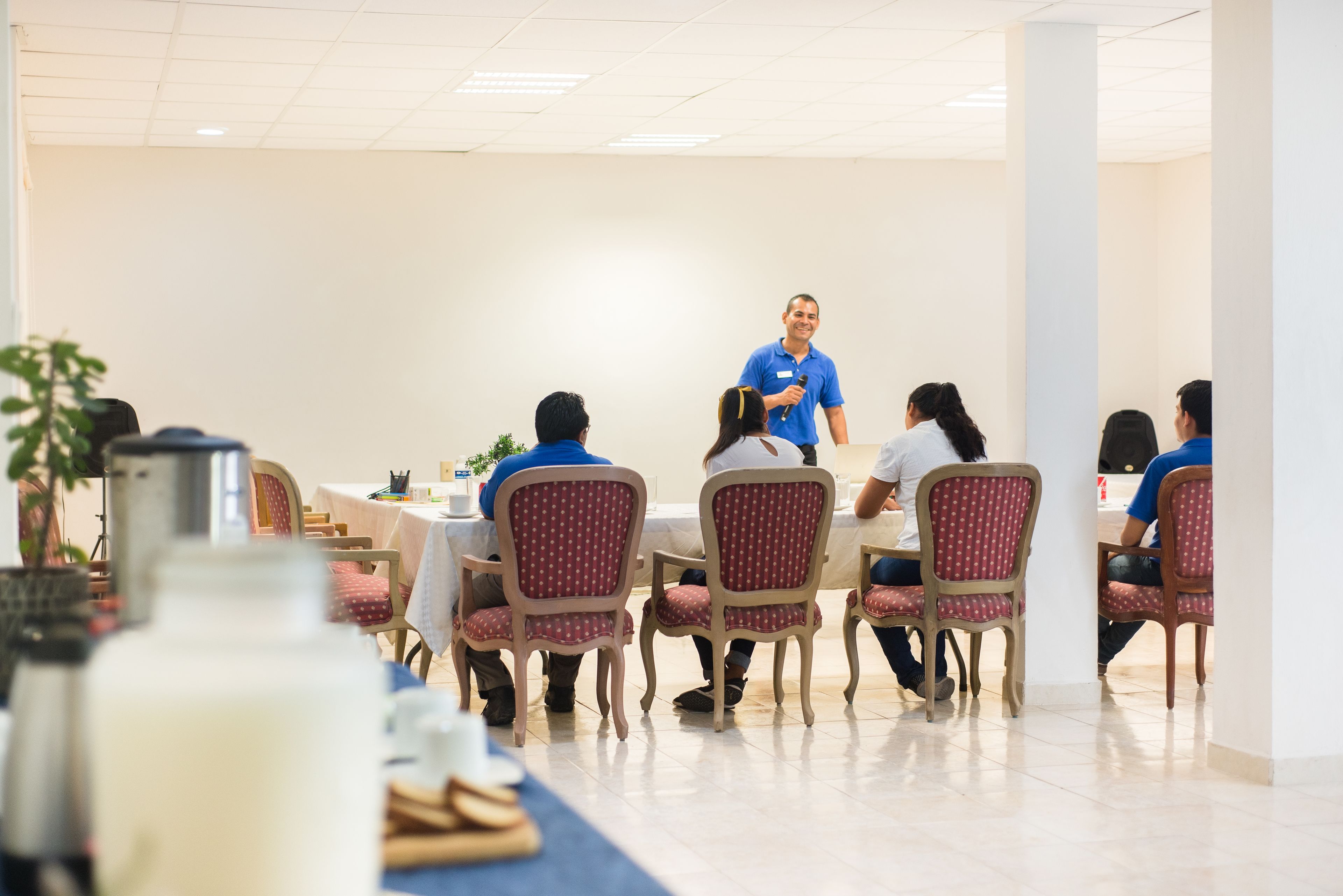 A conference room with a speaker at the front, attendees seated at tables, and a buffet area in the foreground.