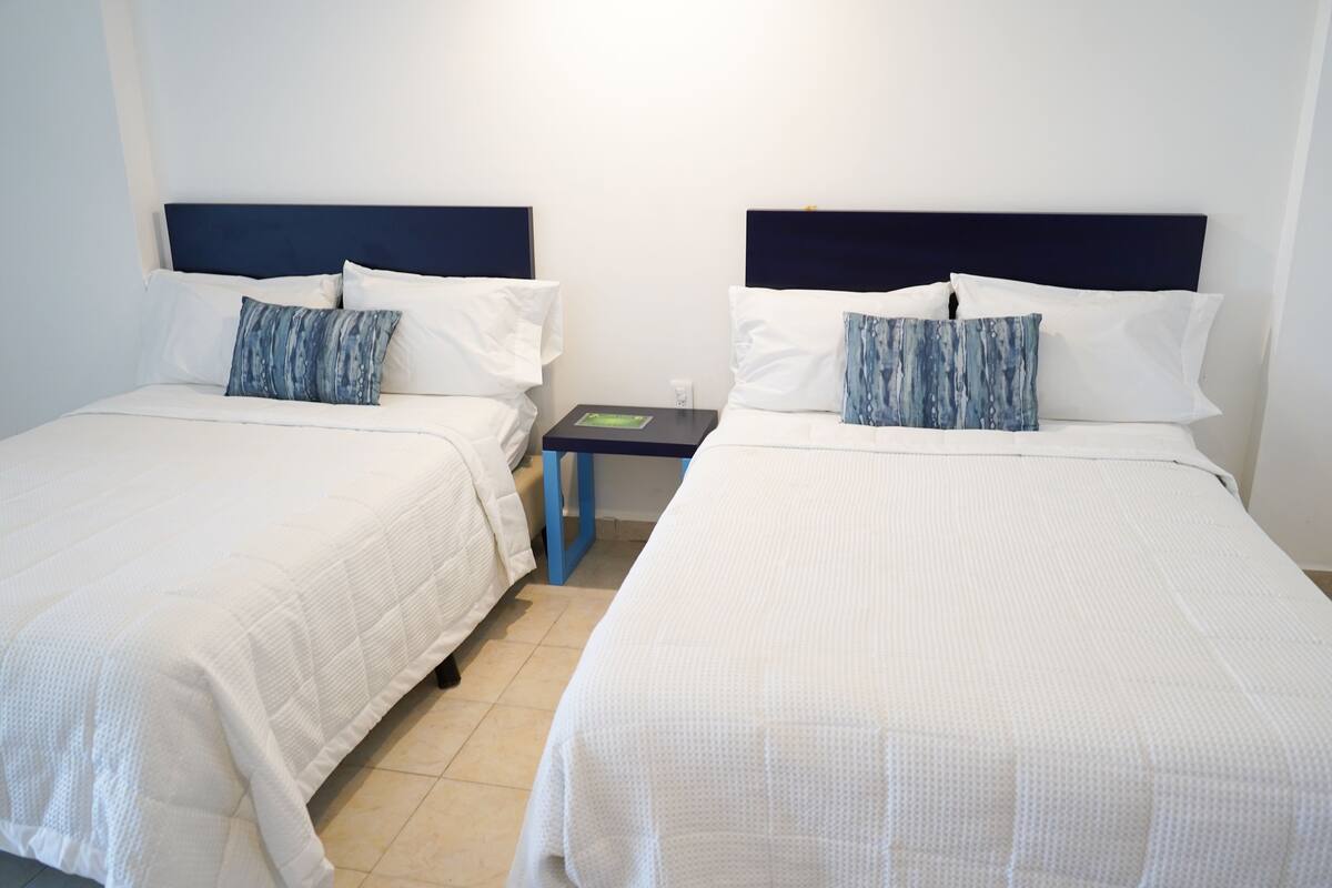 Two single beds with white bedding and blue headboards, a small table with a book, and a tiled floor.