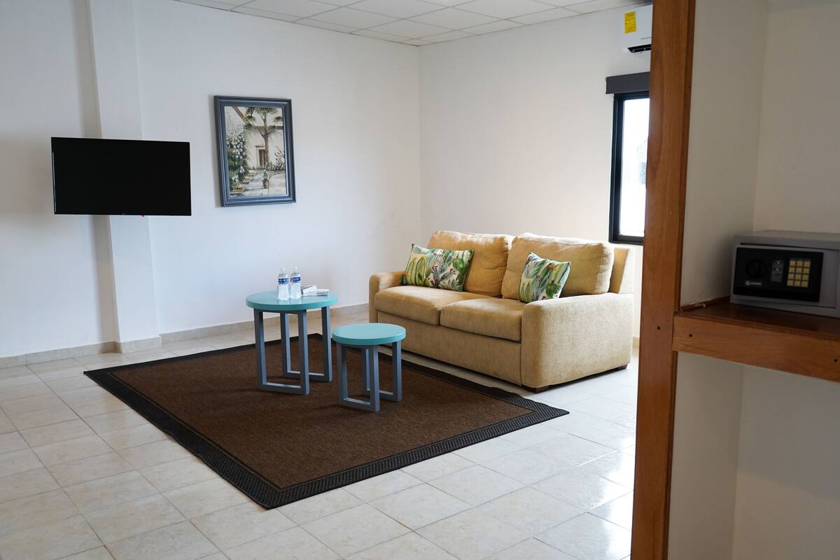 A living room with a beige sofa, two blue stools, a framed picture on the wall, a flat-screen TV, and a safe on a wooden shelf.