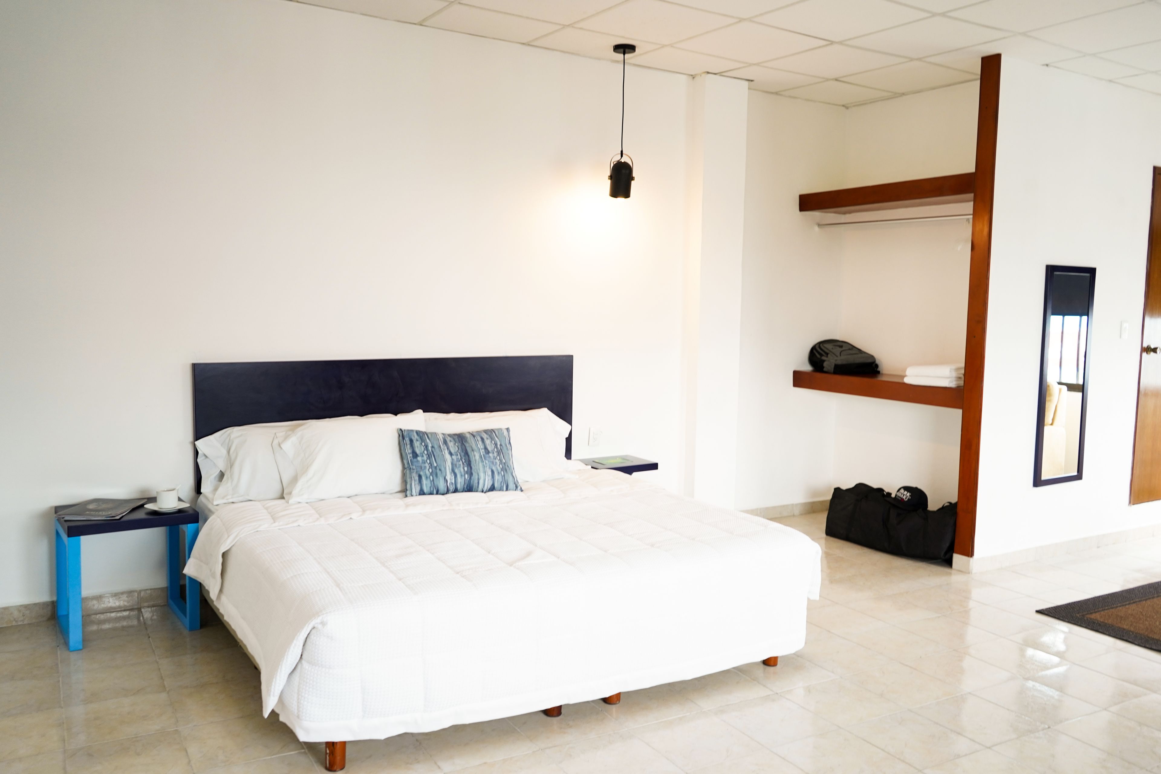 Un cuarto de hotel ordenado con cama, mesitas de noche, lámpara colgante, armario y una maleta en el suelo.