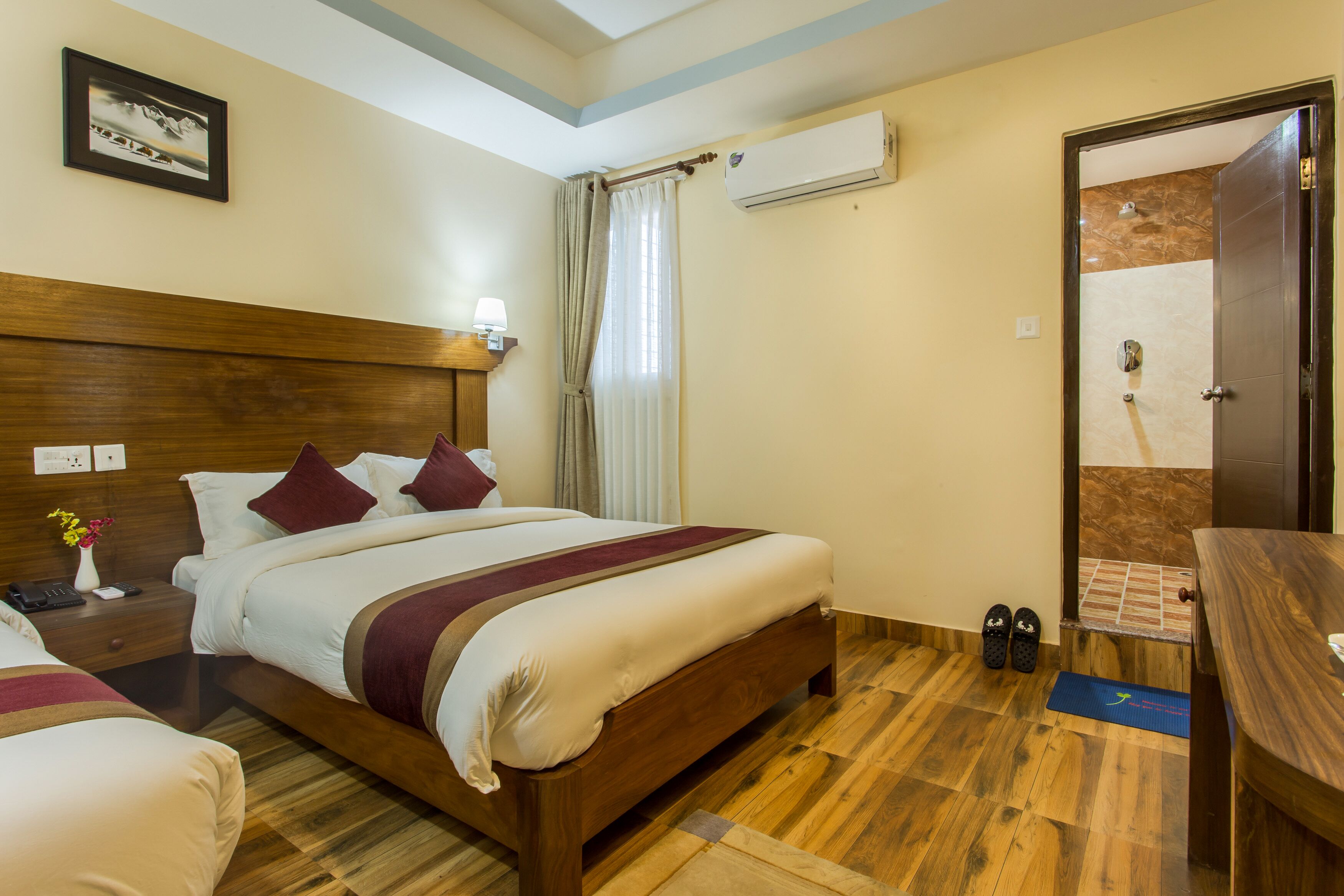 A hotel room with a wooden bed, a nightstand with a lamp, a wall-mounted air conditioner, and a door leading to a bathroom.