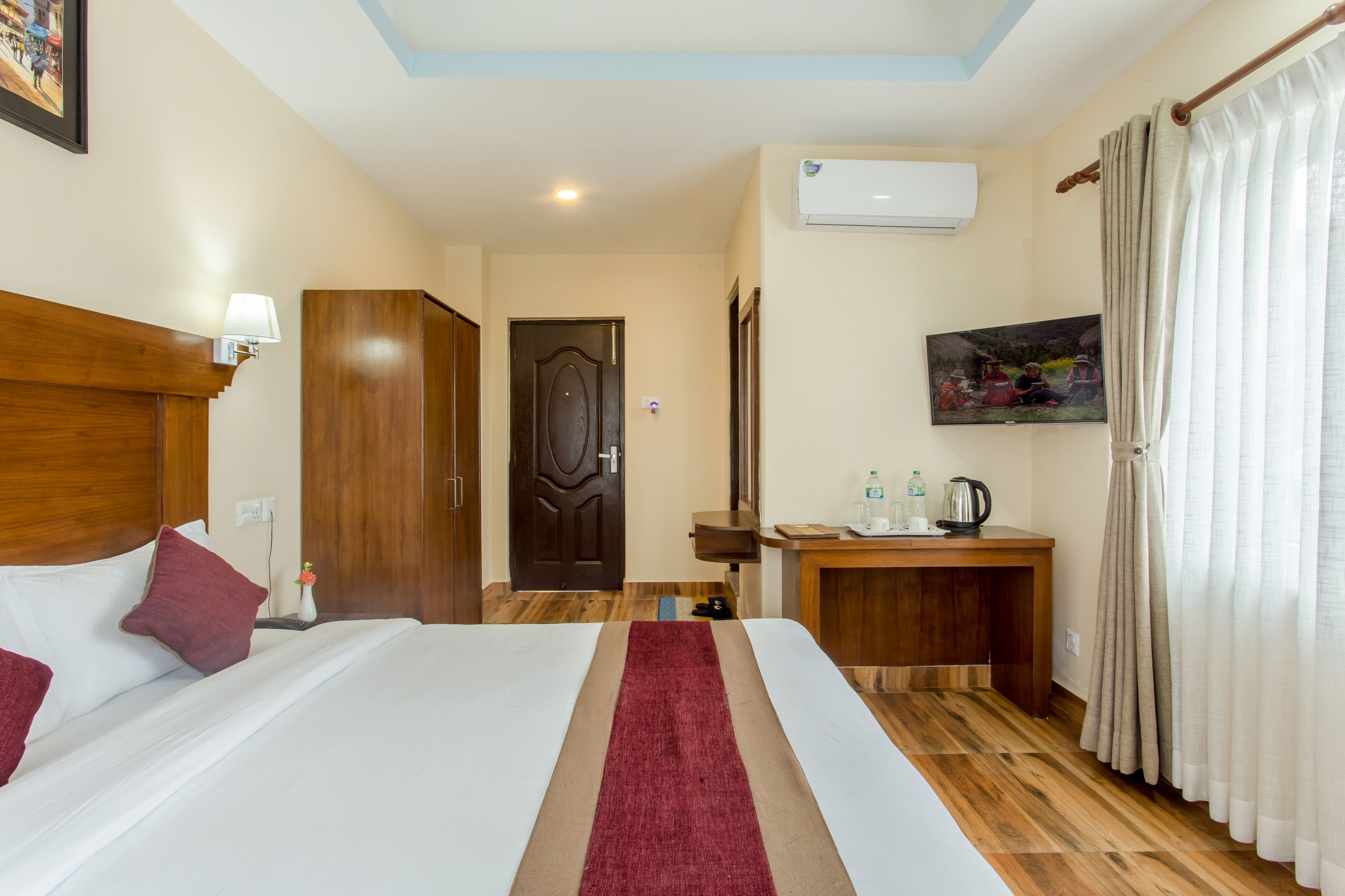 Habitación de hotel con cama, cabecera de madera, escritorio con tetera, una mesita con una botella y un cuadro en la pared.