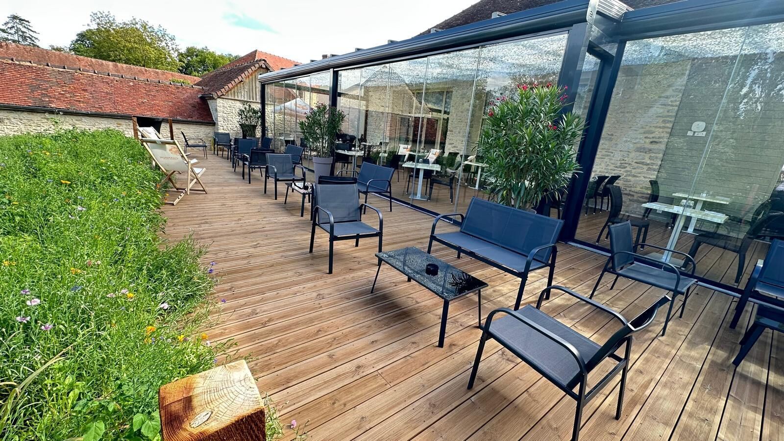 An outdoor patio with wooden flooring, metal chairs, and tables, surrounded by glass walls and potted plants.