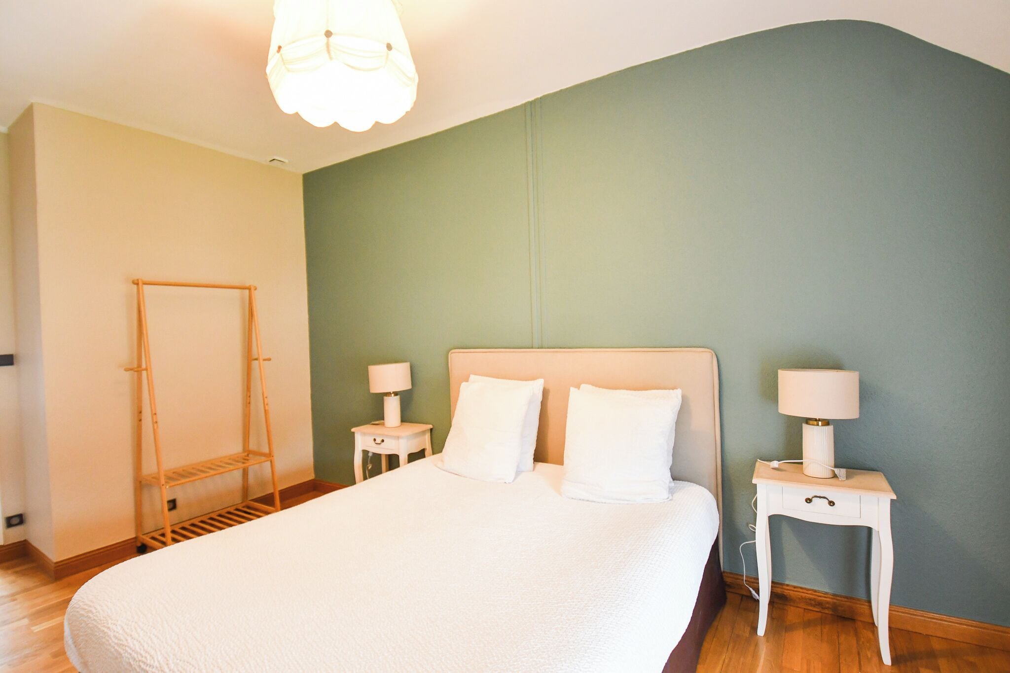 Un lit bien fait, avec du linge de lit blanc, deux tables de chevet avec des lampes et un étendoir en bois adossé à un mur vert.