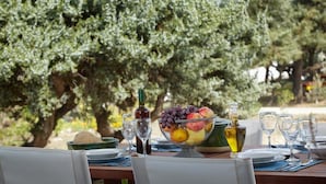 An outdoor dining table set with a bowl of fruit, wine bottles, and glasses, surrounded by olive trees. - Family villa for four with one bathroom, sea view, communal pool, breakfast (Naxos island)