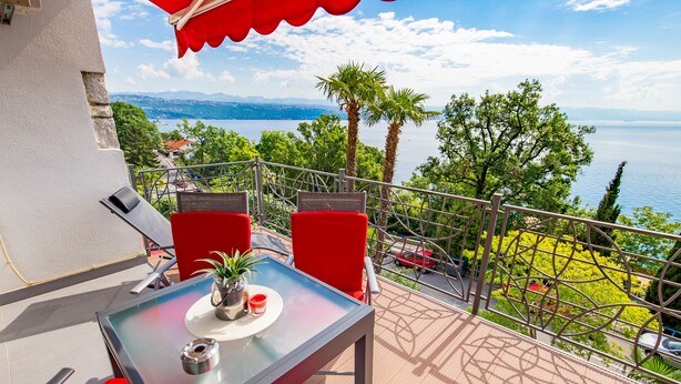 A balcony with a table and chairs, overlooking a lake with trees and a distant mountain range. - Apartment Mia (Opatija)