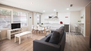 A modern living room with a grey sofa, a white coffee table, and a TV on a white stand. There is a dining area with a white table and chairs, and a kitchen with white cabinets and a stainless steel range hood. - LOFT CELINDA CORDOBA ESTACION (Córdoba)