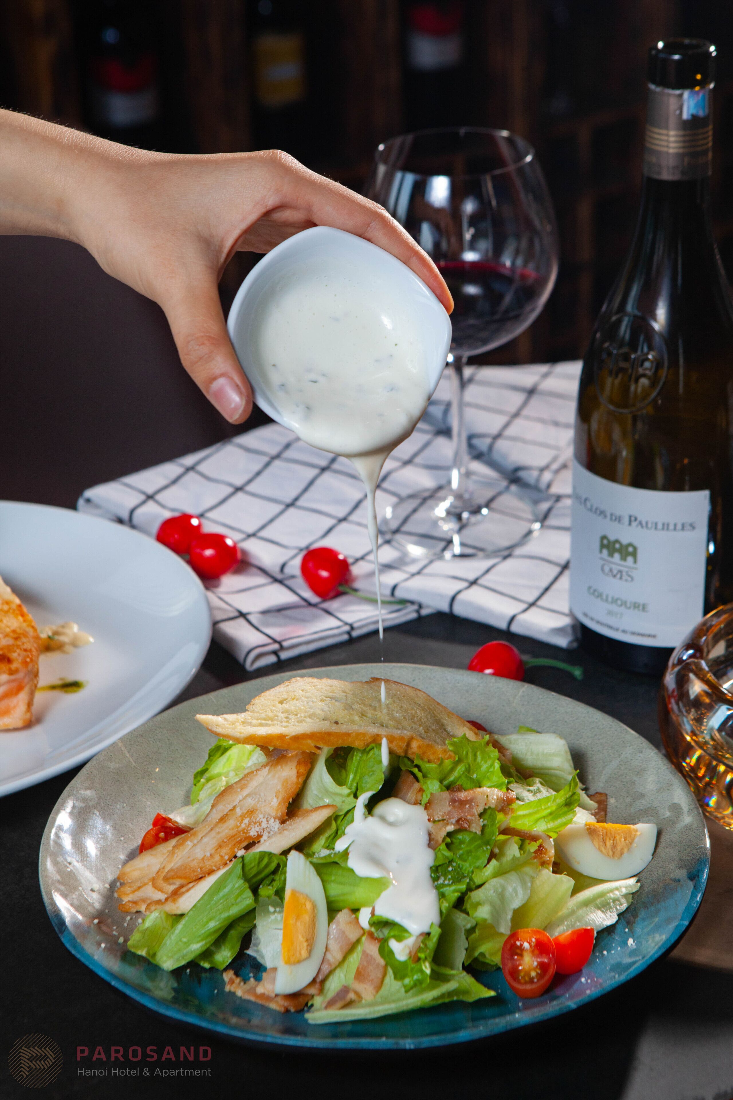 Une main verse une sauce crémeuse sur une salade composée de laitue, de tomates et d’un œuf dur.
